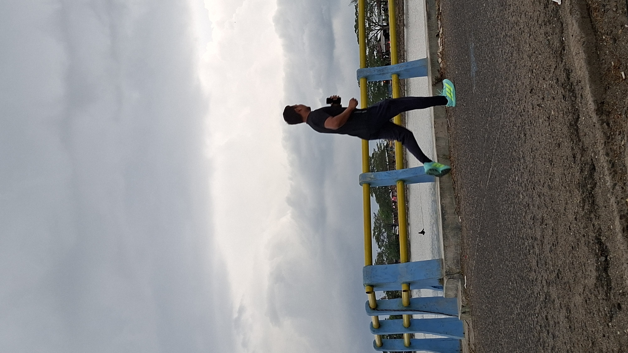 An underexposed, vertical medium shot shows a man jogging across a road. The man, who is facing left and is in profile, is wearing a black t-shirt, black pants, and bright green athletic shoes. He is holding a black object in his right hand. He is running on a dark gray asphalt road. In the background, there is a blue and yellow railing, a body of water, and trees. The sky is overcast and gray. The scene is identifiable as being near the area of Jalan Malikussaleh in Lhokseumawe, Indonesia, based on the distinctive blue and yellow railing style commonly found along the city's waterfront and pedestrian paths. The presence of the body of water and the specific design of the railing are characteristic of the waterfront areas in Lhokseumawe.