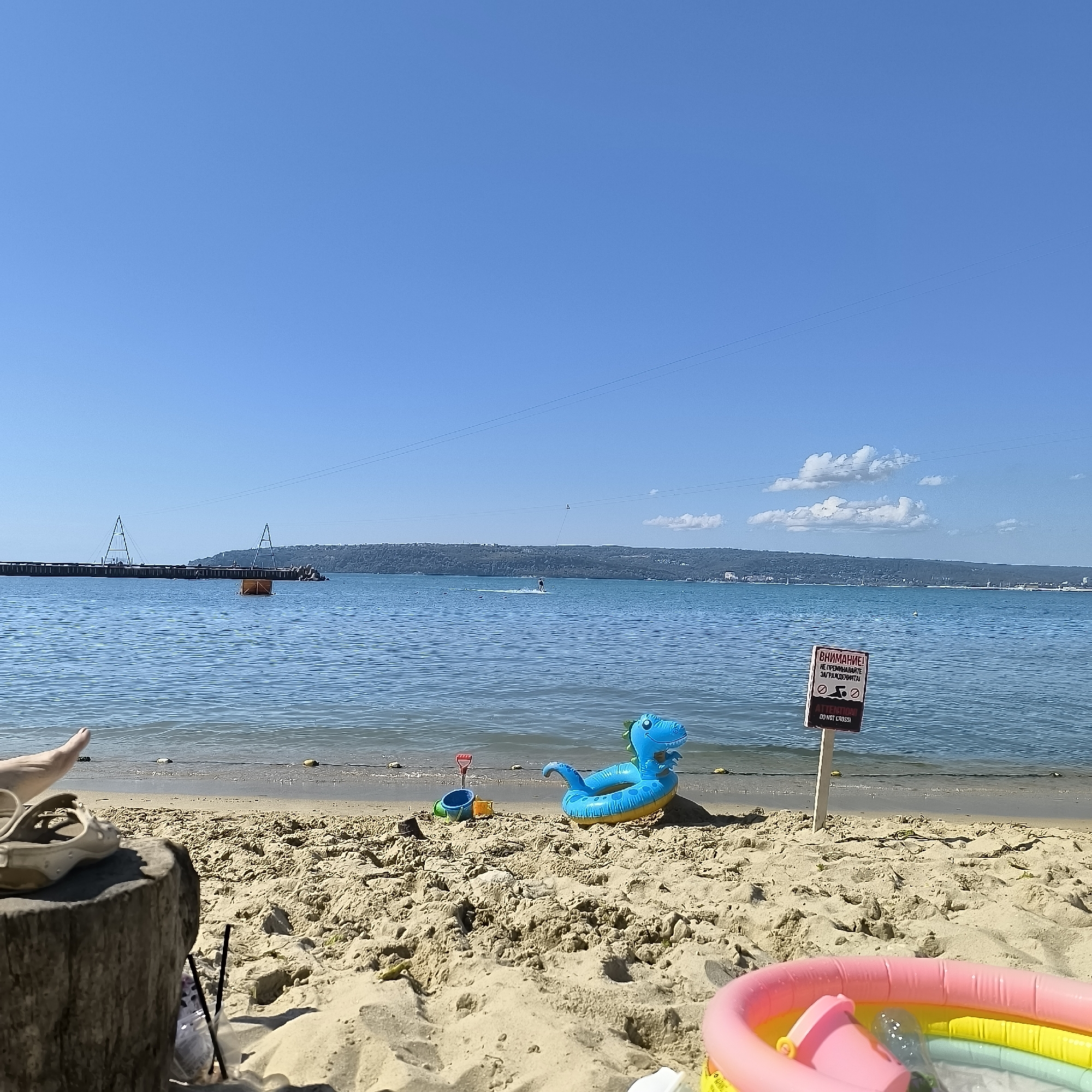 The image depicts a beach scene on a clear, sunny day, likely in Varna, Bulgaria, given the context of a previous query. The foreground is dominated by sand with scattered debris and children's toys, including a blue inflatable dinosaur, a red shovel, and a blue bucket. A pink inflatable pool with yellow and green accents is partially visible in the lower right corner. A pair of beige sandals rests on a weathered wooden stump in the lower left.
The middle ground features the calm, blue sea with gentle waves lapping the shore. A red and white sign with Bulgarian and English text, warning "ATTENTION! DO NOT CROSS!", is planted in the sand near the water's edge. In the water, a person is visible on a stand-up paddleboard, and further out, a long pier with two triangular structures extends into the sea, alongside an orange floating object. A distant coastline with lush green hills and scattered buildings forms the background. The sky is a vibrant, clear blue with a few scattered white clouds. Power lines are visible stretching across the sky.