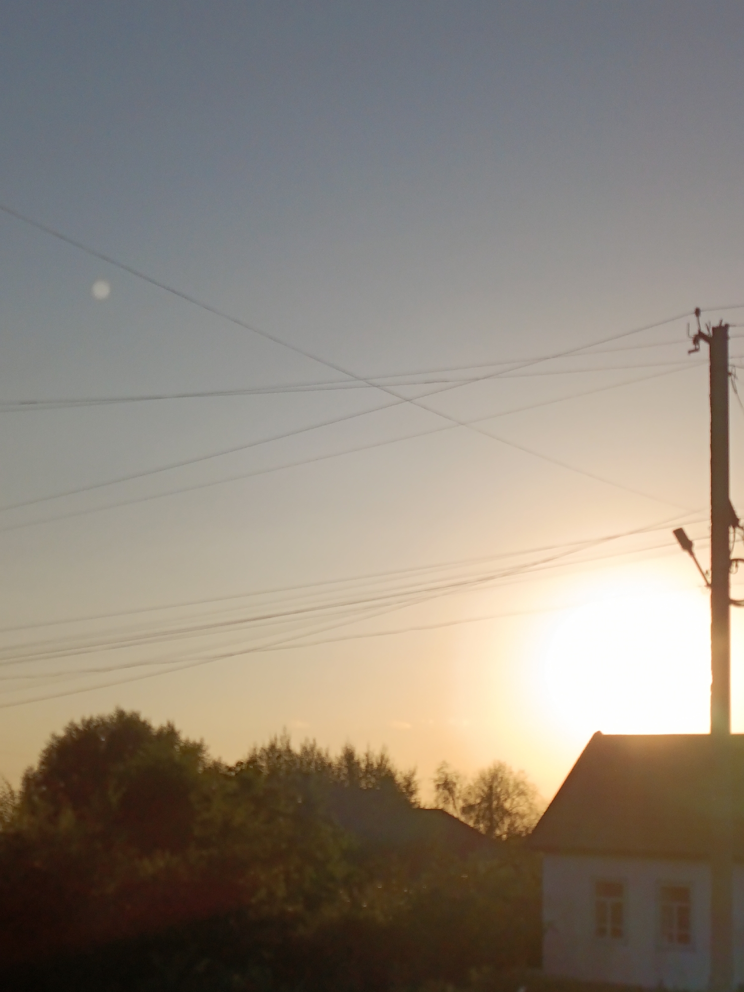 A silhouetted village scene at dusk features a power pole with a streetlight and multiple wires stretching across the sky. In the foreground, the roofline of a building with two windows is visible on the right, partially obscured by trees. The sky transitions from a pale blue to a warm orange hue near the horizon, where the sun is setting. A bright, circular object, possibly the moon, is visible in the upper left quadrant of the sky. The overall atmosphere suggests a rural setting in Kyzyl-Suu, Kyrgyzstan, characterized by natural light and infrastructure.