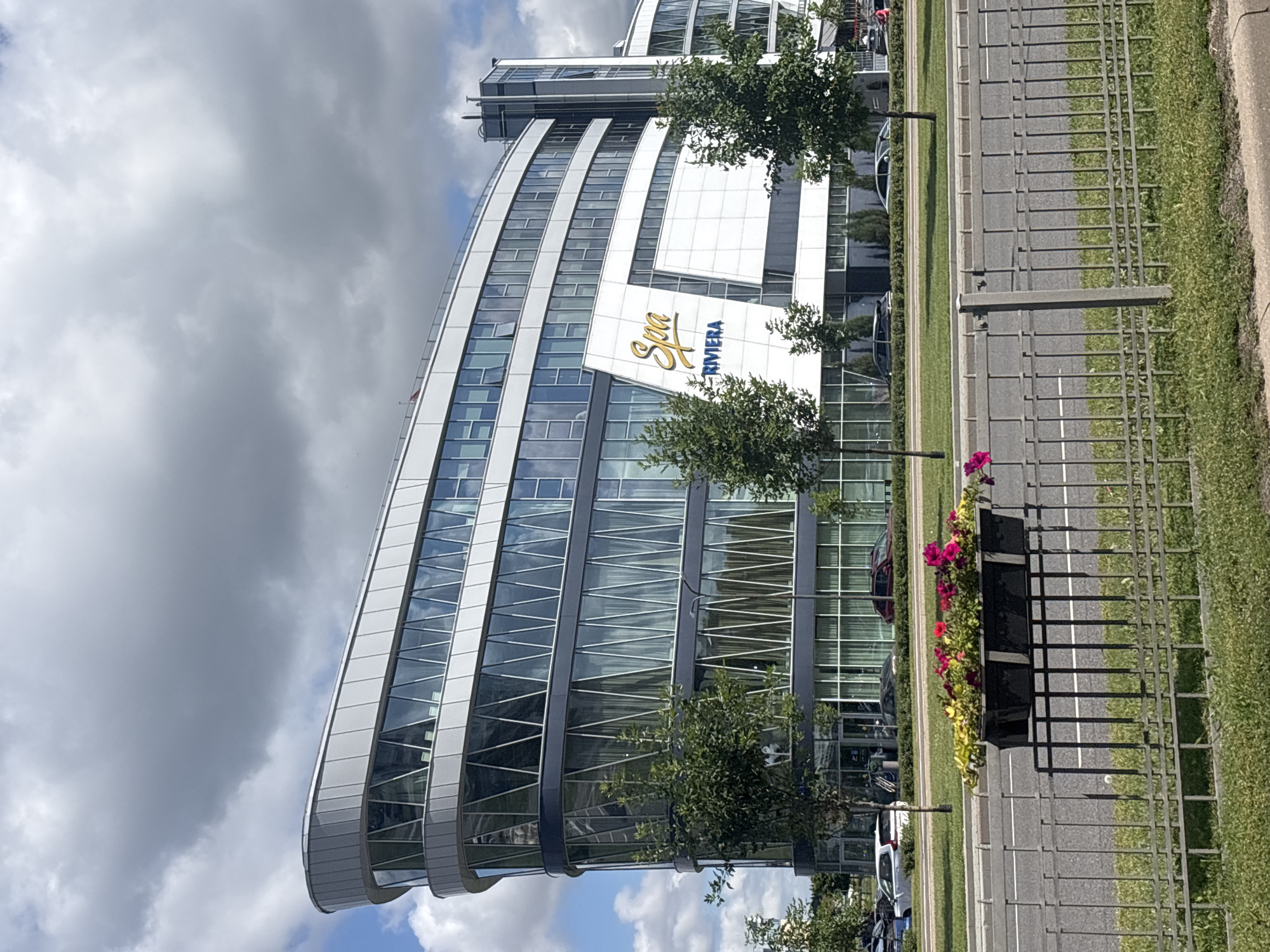 The image shows the modern architecture of the Spa Riviera building located in Minsk, Belarus. The building is characterized by its curved glass facade and white accents. A large sign with "Spa" in yellow script and "RIVIERA" in blue lettering is prominently displayed on the building. In front of the building, there are trees and a street with cars. A fence separates the street from a grassy area with a planter box filled with colorful flowers. The sky is partly cloudy.