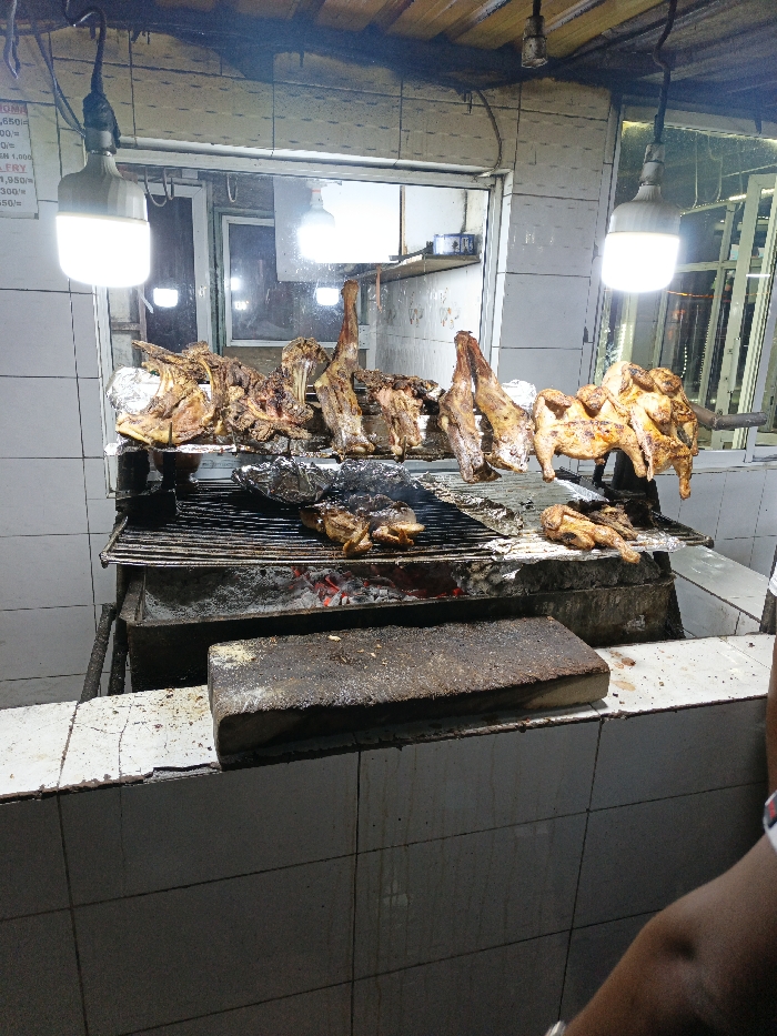The image depicts an active indoor commercial grilling station, typical of a Nyama Choma (grilled meat) eatery in Nairobi, Kenya.
In the center, a large, rectangular charcoal grill contains visible red embers beneath a metal grate. Numerous pieces of roasted meat are present: five large cuts, consistent with goat or lamb, hang or rest on the grate, alongside multiple whole or half chickens. Some items appear partially cooked, while others are fully roasted. Several objects wrapped in aluminum foil are also on the grill.
The background features white, stained tiled walls, with a window or opening revealing additional interior lighting. Two bright white LED lamps illuminate the scene from above. A partial white sign on the far left wall displays black text, including price indications and food items such as "CHICKEN," "FULL," and "HALF."
A low, white-tiled counter, showing surface cracks and general wear, occupies the foreground. It features a large, dark, rectangular wooden cutting board. A partial human arm is visible in the lower right quadrant of the frame. The scene indicates ongoing food preparation and sale in a utilitarian, commercial kitchen environment.