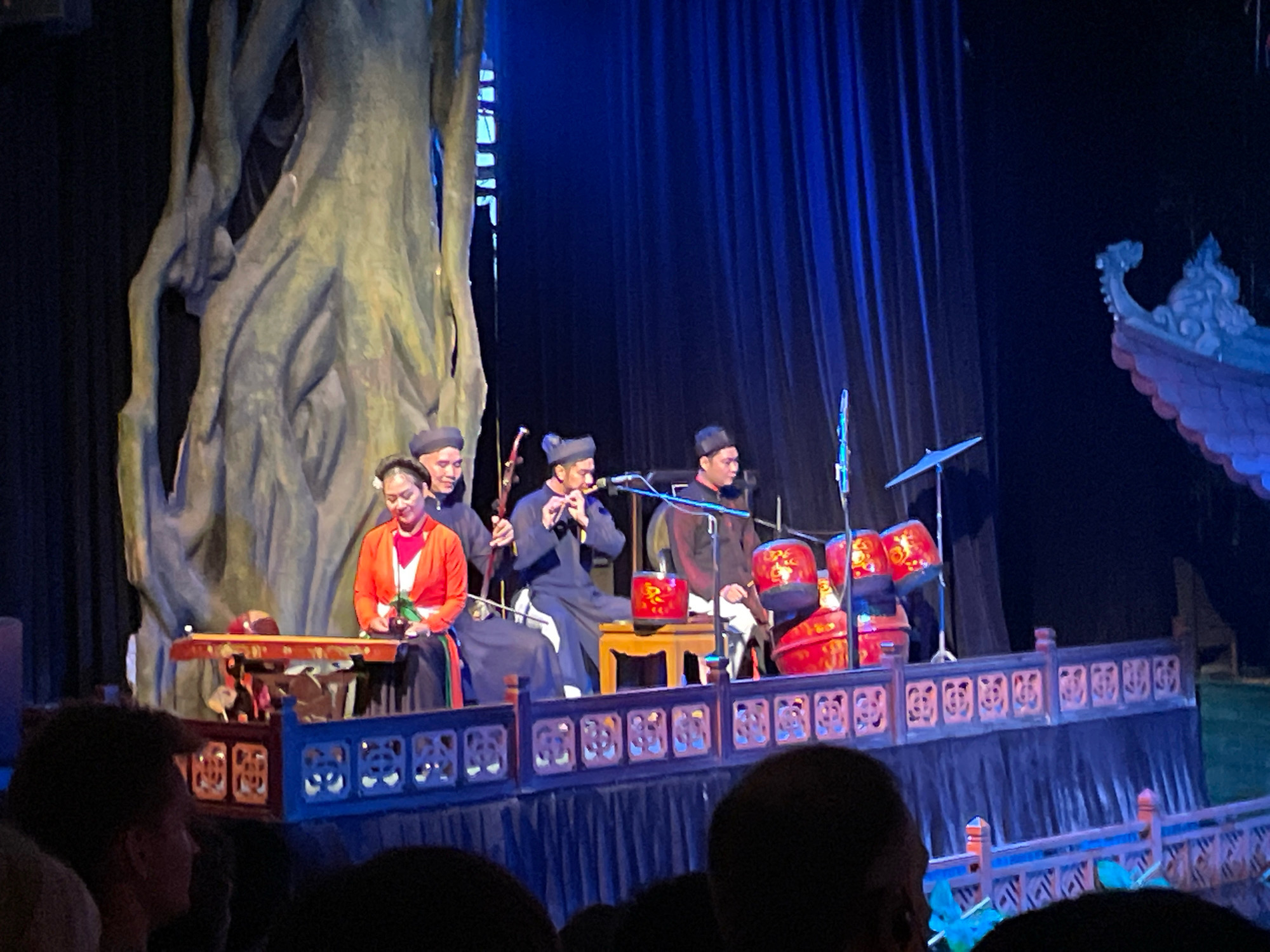 This image captures a scene at the Thang Long Water Puppet Theatre in Hanoi, Vietnam. On a stage, several performers are actively engaged in a traditional water puppet show. In the foreground, a woman in a vibrant orange and red outfit sits playing a stringed instrument. To her right, a man in traditional attire plays a flute, his instrument positioned near a microphone. Further right, another man in similar clothing is seated, possibly another musician. Behind them, a large, abstract tree-like structure serves as a backdrop, its gnarled roots and trunk dominating the left side of the frame. The stage is decorated with a blue and red railing adorned with intricate carvings. Red drums with gold embellishments are visible on the right side of the stage. The background features dark blue curtains. The lighting is dramatic, with blue stage lights illuminating the performers and the set. In the lower part of the image, the backs of audience members are visible, indicating the presence of spectators watching the performance.