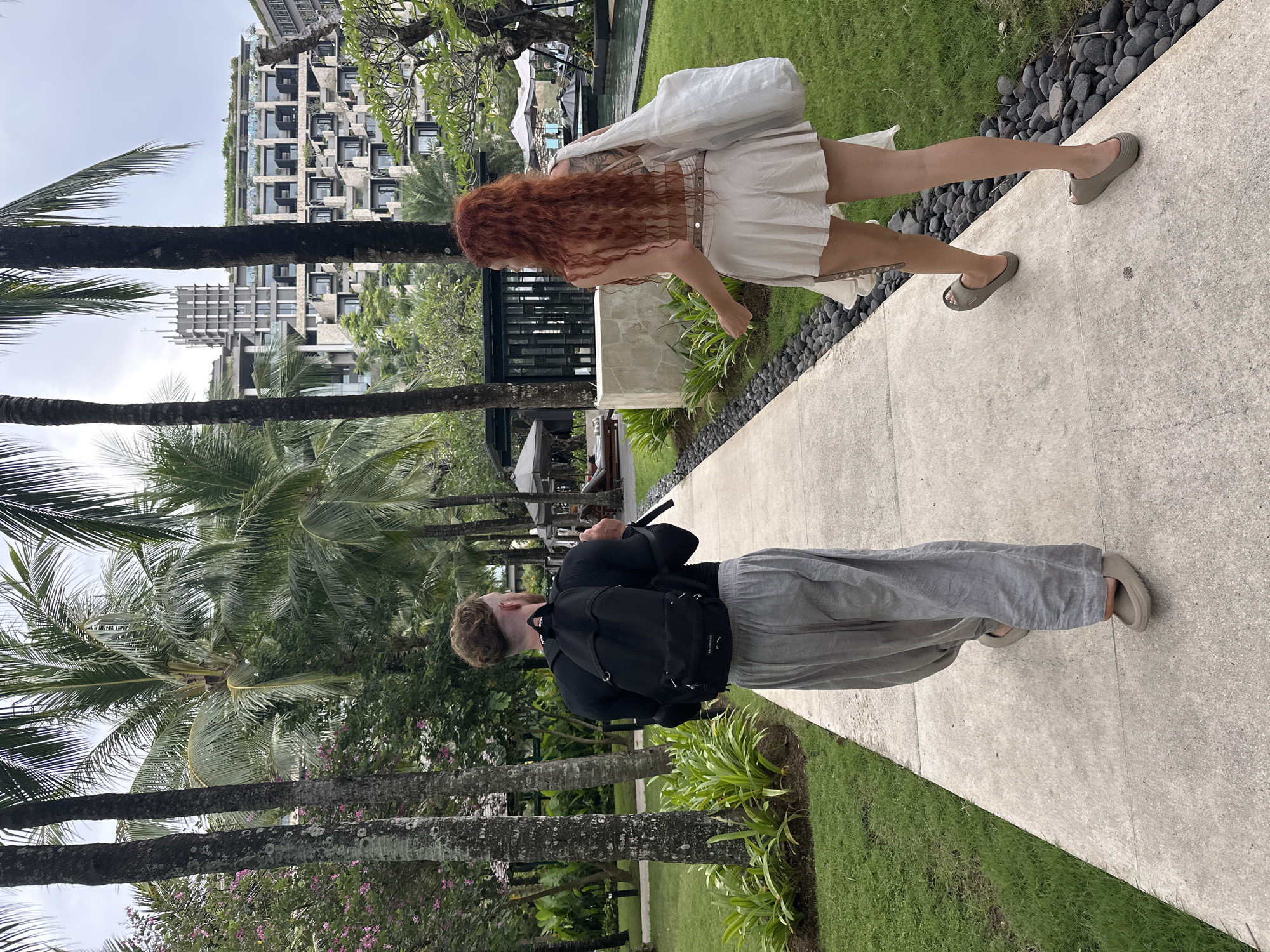 In Nusa Dua, Indonesia, a photo captures two individuals on a paved path. A person with long red hair, wearing a white dress and carrying a white bag, walks slightly ahead. Behind, another person with short blond hair wears a black shirt, gray pants, and a black backpack. Palm trees line the path, with a modern building visible in the background. The scene includes a mix of pavement, grass, and tropical foliage.