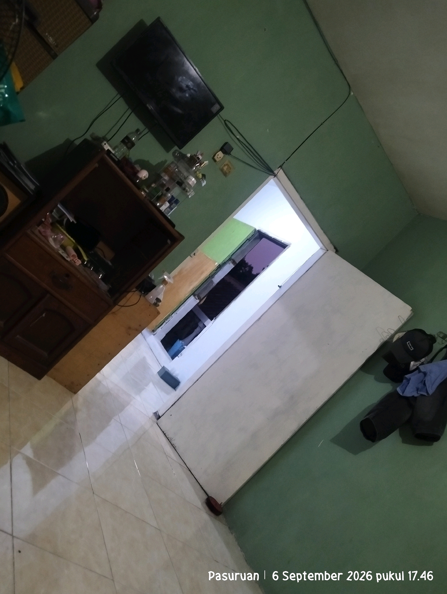In Warung Dowo, Indonesia, the image depicts an interior scene, likely a residential room, characterized by a slightly disheveled state. A black flat-screen television is mounted on a pale green wall. Below it, a dark wooden cabinet with drawers and shelves is partially visible. On the shelves, various items are present, including bottles and other indistinct objects. To the right of the cabinet, a light-colored wooden structure extends, supporting a clear plastic bottle.
The room features a doorway that is open, revealing a brightly lit interior with a purple-tinted window or screen. A large, white, flat surface, possibly a door or a partition, dominates the lower right portion of the image. Scattered on the floor near this surface are clothing items, including a black baseball cap, dark trousers, and a blue shirt. The floor itself is tiled with a light-colored, square pattern, and appears somewhat reflective. The overall lighting is dim, with the bright light from the open doorway being a significant light source. The timestamp "Pasuruan | 6 September 2026 pukul 17.46" is overlaid on the lower right corner of the image, indicating the location and time of capture.