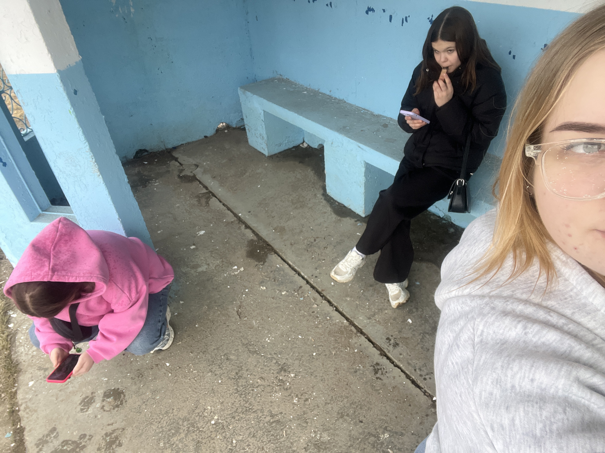 Three young women are in what appears to be a bus shelter. One woman is crouching and looking at a red smartphone. A second woman is sitting on a concrete bench, holding a purple smartphone, and eating. The third woman is taking a selfie. The bus shelter is made of concrete painted blue and white. The ground is concrete and has some debris scattered on it. The location is Chaikovsky, Russia.