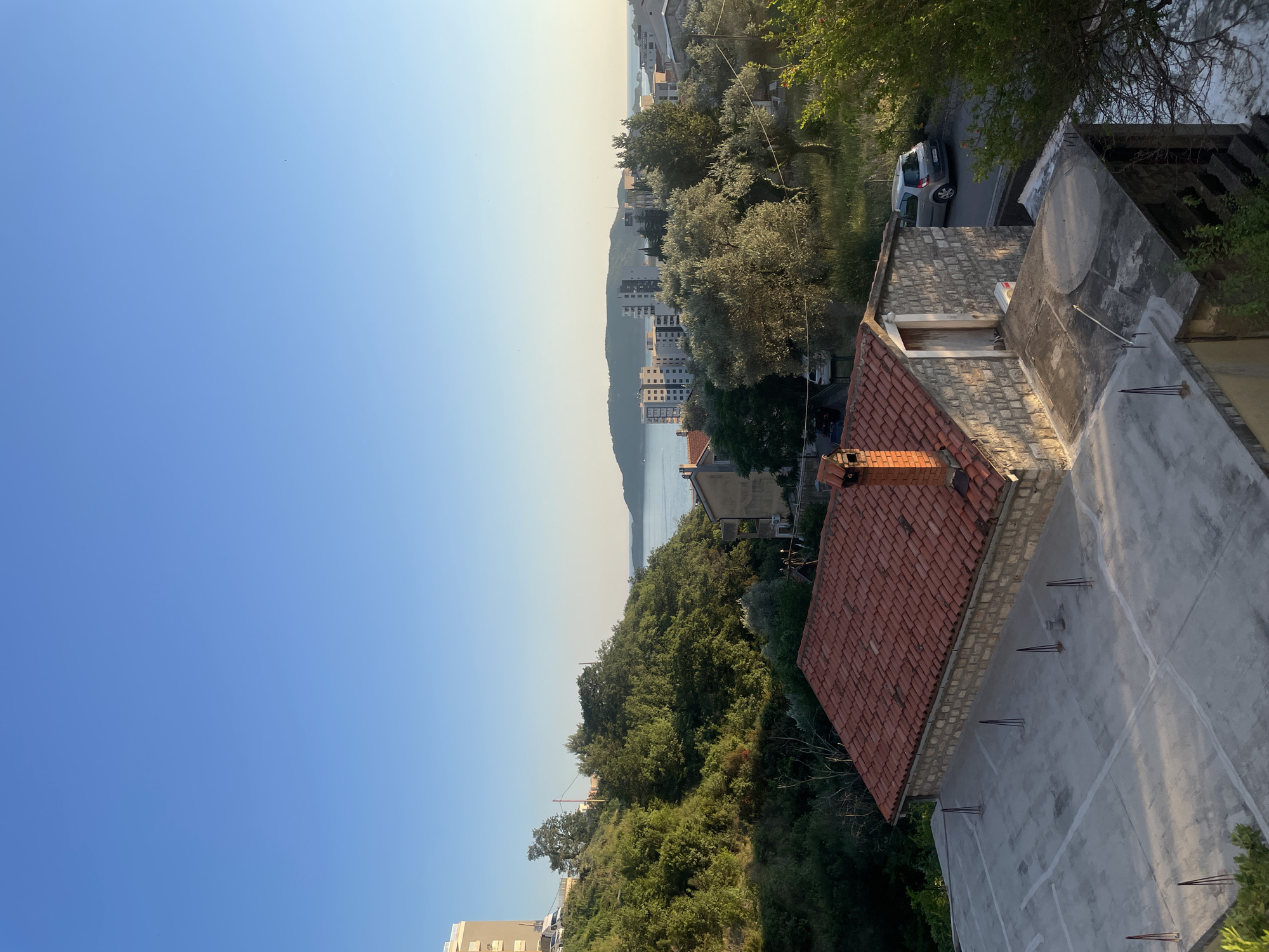 The image captures a daytime view overlooking the coastal city of Rafailovići, Montenegro. In the foreground, a partially constructed flat concrete rooftop is visible, with several metal rods protruding from it. Adjacent to the rooftop is a small, stone-built structure with a red-tiled roof and a brick chimney. A silver hatchback car is parked on a paved road to the right of this structure.
The middle ground is dominated by lush green trees covering a hillside, with a few buildings interspersed among them. Further in the distance, the cityscape of Rafailovići unfolds, characterized by multi-story apartment buildings and other structures. Beyond the city, a body of calm water, likely the Adriatic Sea, is visible, with a hilly island in the far distance. The sky is a clear, pale blue, indicating fair weather. The lighting suggests it is either early morning or late afternoon, given the warm, soft illumination.