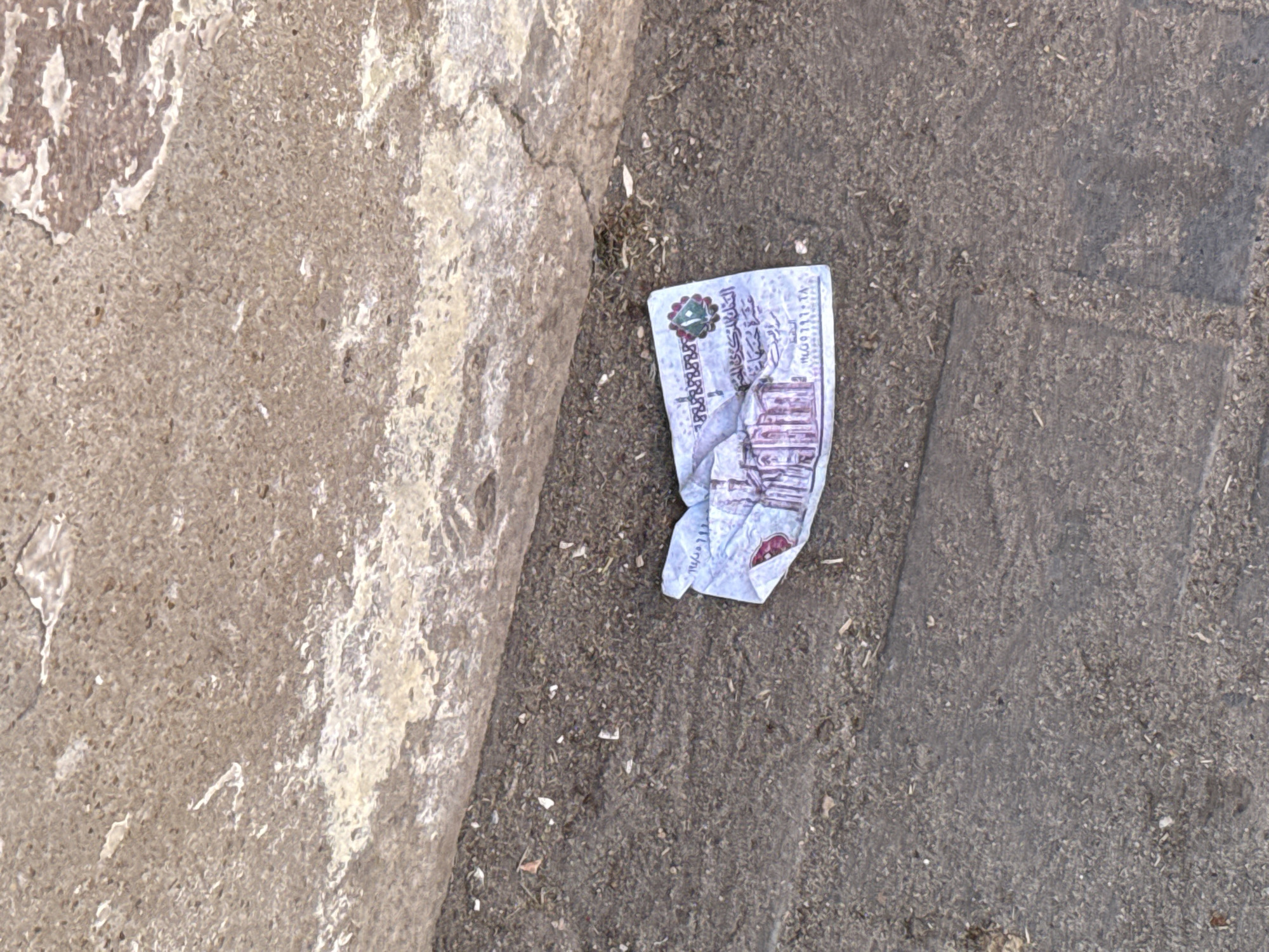 A crumpled Egyptian ten-pound note lies on a dirty, textured surface, possibly a sidewalk or road. The note is light blue with intricate designs and Arabic writing. In the background, a rough, beige concrete structure runs along the top of the frame. The surface beneath the note is dark and appears to be composed of small pebbles and dirt, with scattered debris. The scene is consistent with an outdoor environment in a city like Giza, Egypt, where such currency and urban textures are common.