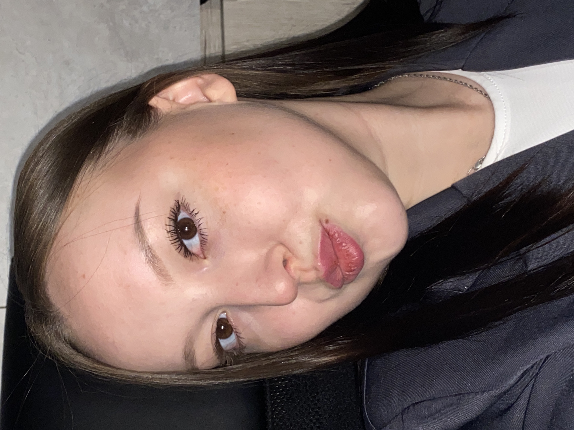 A close-up portrait of a young woman, taken indoors, is depicted in the image. She has light skin with faint freckles, long dark brown hair, and brown eyes with visible mascara. Her hair is styled with a side parting, framing her face. She is wearing a dark-colored jacket or blazer over a white top, with a thin silver chain necklace visible around her neck.
The woman's face is central to the composition, and she is looking upwards and slightly to the side with a direct gaze. Her lips are distinctly pursed, forming a "duck face" expression, which conveys a playful or pouty emotion. The lighting, possibly artificial, illuminates her face, highlighting features such as her forehead, nose, and cheeks. The background is indistinct, dark, and appears to be a plain indoor surface. There are no discernible location cues, specific time of day indications, weather details, or visible text within the image. The woman is actively posing for the photograph.