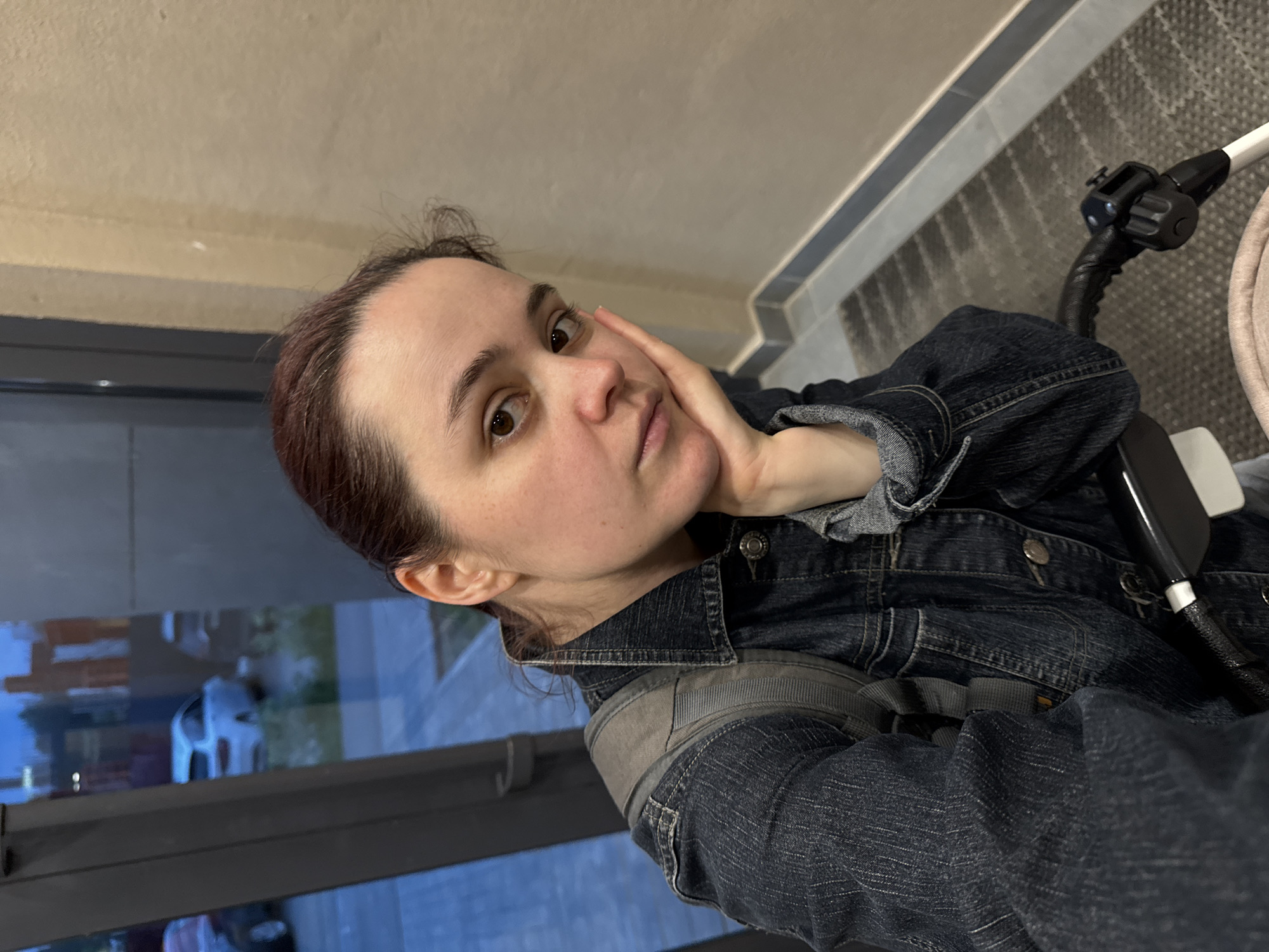 A woman with brown hair is seated in a stroller, wearing a denim jacket and backpack. She is resting her head on her hand, looking at the camera. Outside the window, there is a glimpse of a building and a car, suggesting an urban environment. Without further information, pinpointing the location within Izhevsk, Russia is impossible.