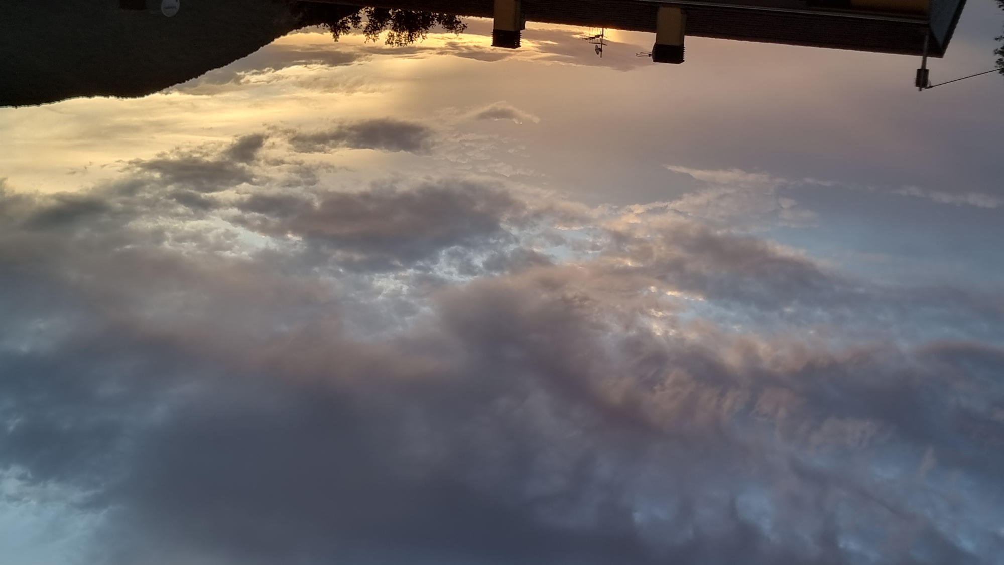 The image shows a dramatic sunset over the town of Sant Joan Les Fonts, Spain. The sky is filled with dark, heavy clouds tinged with pink and orange from the setting sun. Below the clouds, a gradient of light from pale yellow to a soft lavender stretches across the horizon. Silhouetted against this colorful sky are the rooftops of houses, some with chimneys, and a distant, dark hillside. The overall impression is one of a peaceful evening with a striking natural phenomenon.