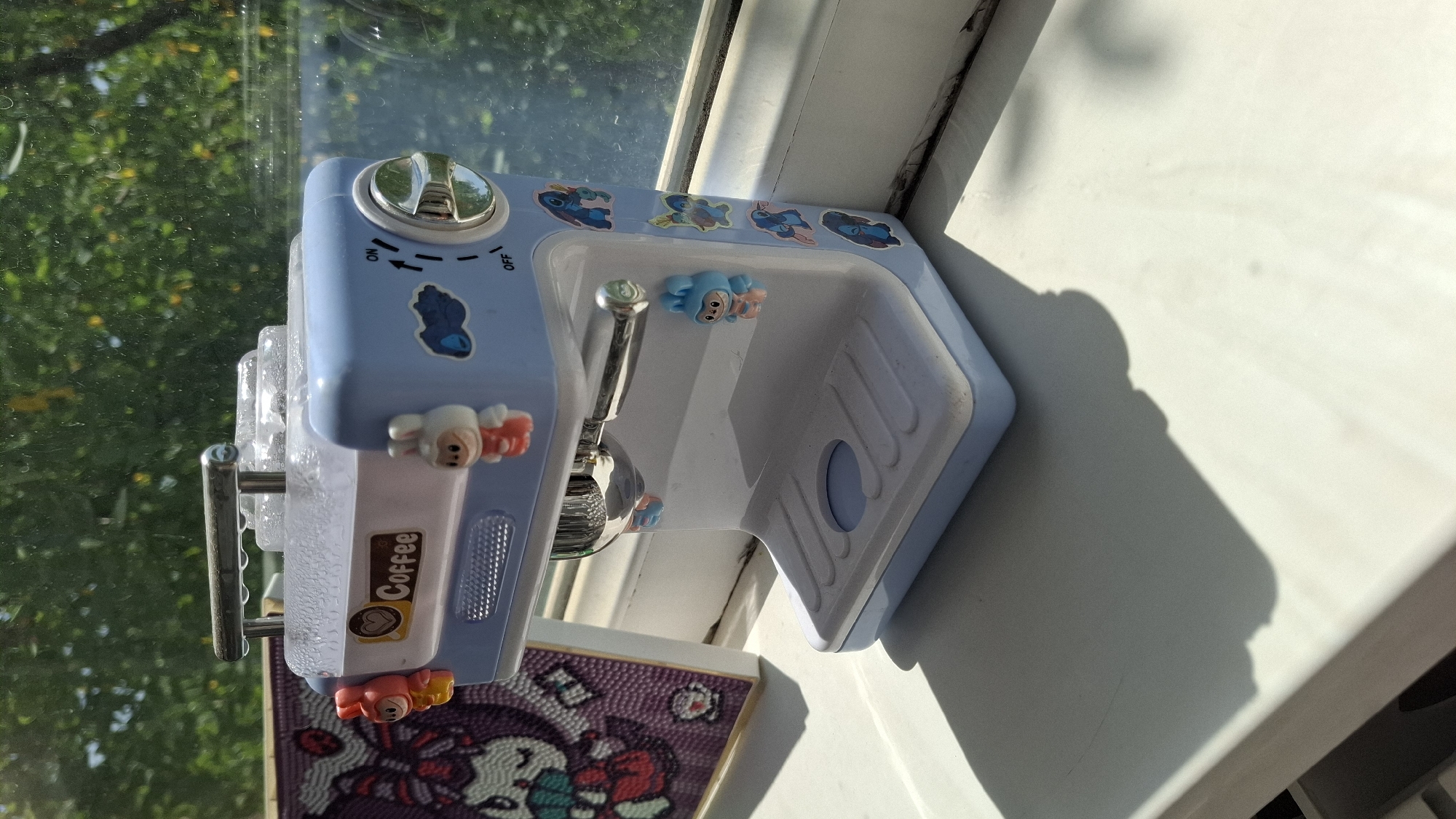 A light blue toy espresso machine with cartoon character stickers sits on a white windowsill. The machine has a chrome handle on top and a dial on the side that reads "ON" and "OFF." There are small, colorful figurines of cartoon characters attached to the machine, including bunnies and what appear to be Stitch from Lilo & Stitch. To the left of the espresso machine is a framed artwork, possibly a diamond painting, depicting a character with large eyes and a bow. The background shows green foliage and trees, suggesting an outdoor view. The scene is illuminated by sunlight, casting shadows on the windowsill. The image was taken in Shymkent, Kazakhstan.
