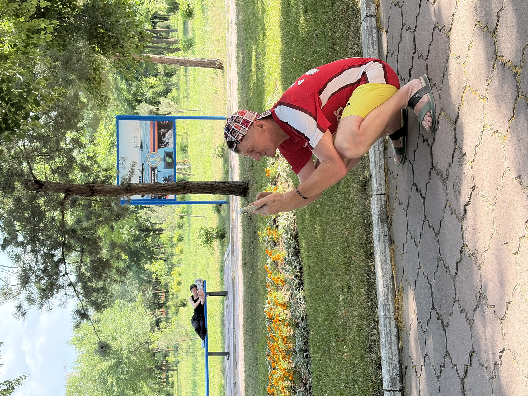 In an outdoor park setting, a man wearing a red and white shirt, yellow shorts, and a patterned baseball cap is squatting on a paver pathway and holding a smartphone. He is looking towards the left, seemingly taking a photograph or video. To his left, on a blue park bench, a person in black clothing is lying down, resting their head on a pillow. Behind the bench, a flower bed with orange flowers is visible, bordered by green grass. In the background, trees and a blue information board with images of ships are present. The sun casts shadows on the ground, indicating it is daytime. The overall environment suggests a public park.