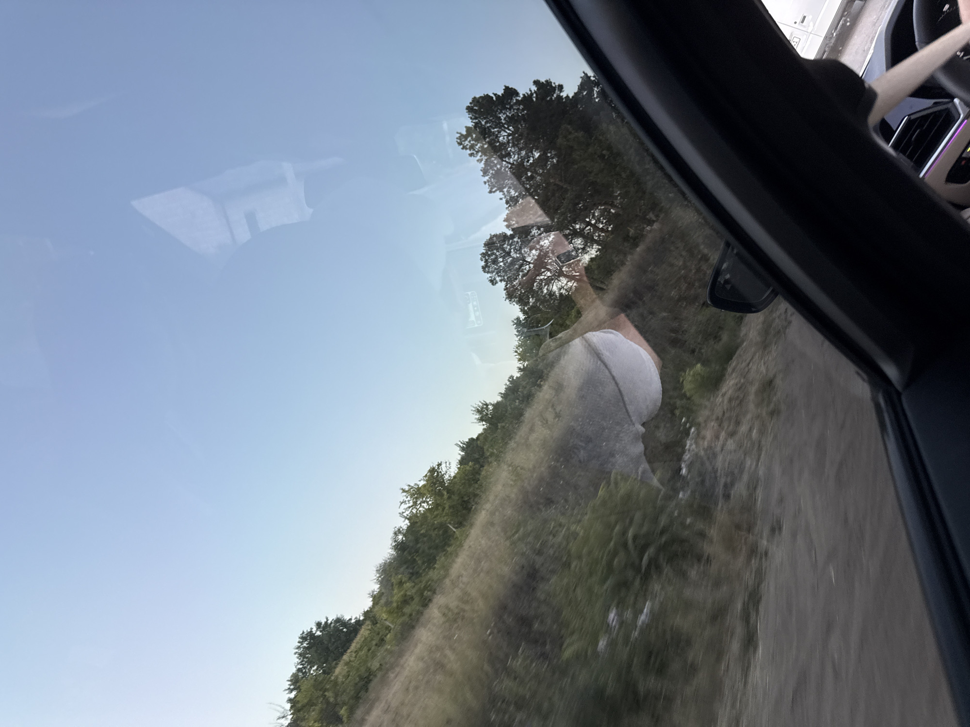 The image is taken from inside a car, looking out through the windshield. The perspective suggests the car is moving. The scene outside the car depicts a rural landscape with a dirt road on the left and a hillside covered in dry grass and green bushes on the right. Several trees are visible on the hillside and in the distance. The sky is a clear, pale blue, indicating daytime. There are reflections on the windshield, including a faint image of what appears to be a person's torso wearing a light-colored shirt or top, and a reflection of the car's interior, including a portion of the dashboard and steering wheel. The overall impression is a scenic drive through a sparsely vegetated, rural area, possibly in Sărata-Mereșeni, Moldova.