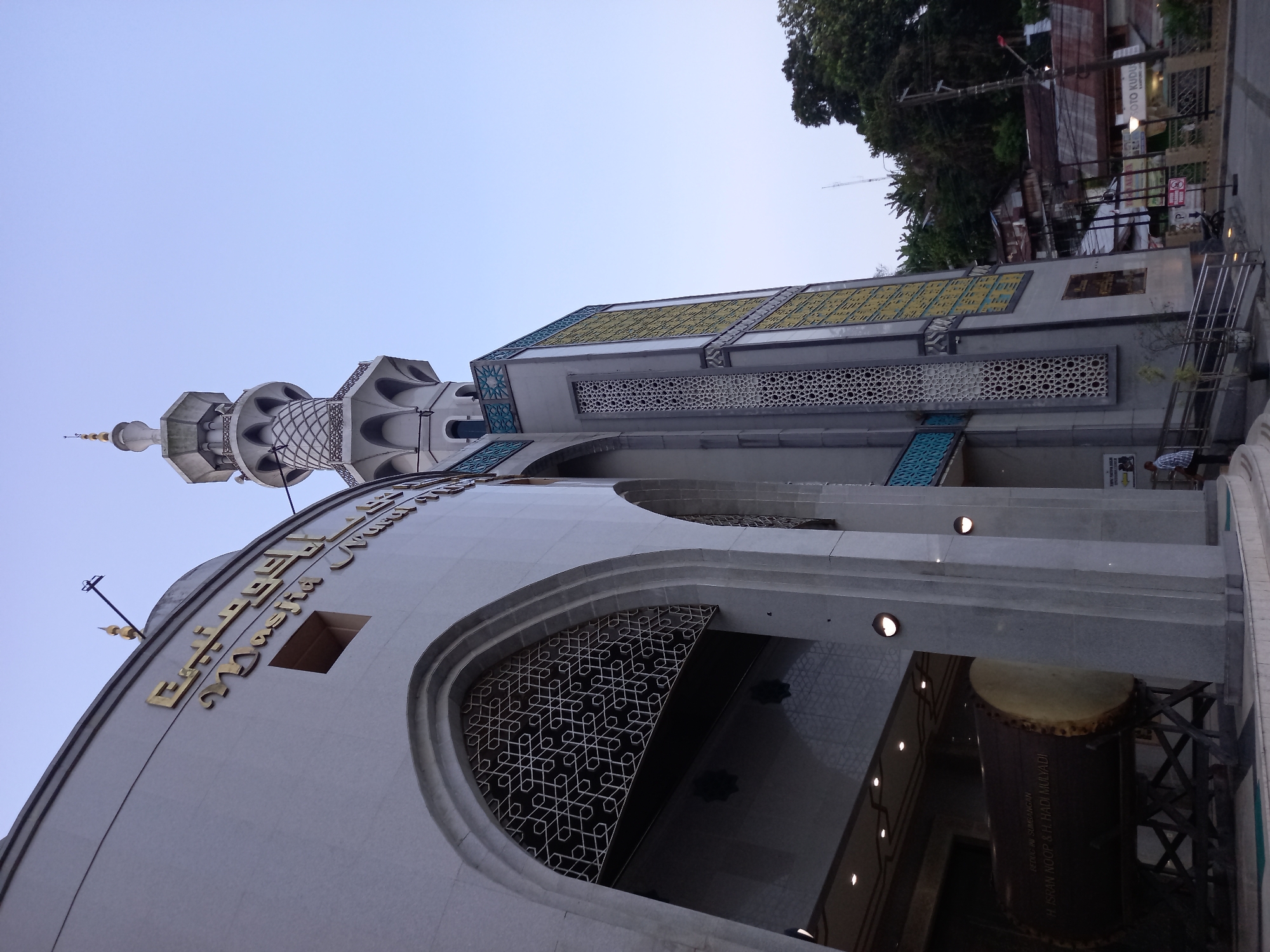 A low-angle, wide shot captures the exterior of a mosque in Samarinda, Indonesia, with a clear sky in the background. The mosque's facade is predominantly light gray, with ornate architectural details. To the left, a large, arched entryway features intricate white geometric patterns. In front of the entryway, a very large, wooden drum, likely a bedug, rests on a wooden stand, with Arabic calligraphy visible on its surface. To the right, a tall, slender minaret rises, adorned with decorative latticework and a finial at its peak. The minaret is connected to a rectangular section of the building featuring yellow and white geometric paneling. A few trees and a hillside with buildings can be seen behind the mosque. A single person, dressed in a light-colored shirt and dark pants, stands near the entrance, facing away from the camera. The overall scene suggests an early morning or late afternoon setting due to the soft lighting.