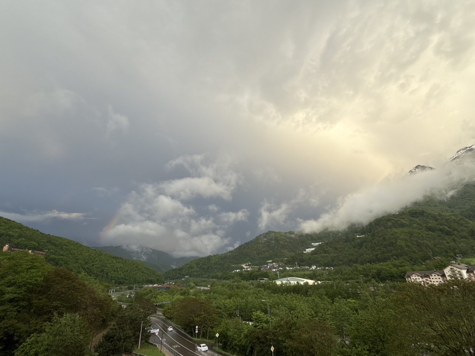 The image depicts a scenic view in Krasnaya Polyana, Russia, under a dramatic sky. A rainbow is visible on the left, arching over green, mountainous terrain. The right side of the image is dominated by mountain peaks partially obscured by low-hanging clouds. A road with a couple of white cars runs through the lower portion of the frame, lined with trees. Buildings are scattered throughout the scene, nestled among the greenery, indicating a settled area within the mountainous landscape. The sky ranges from dark, stormy clouds on the left to a brighter, sunlit area on the right.