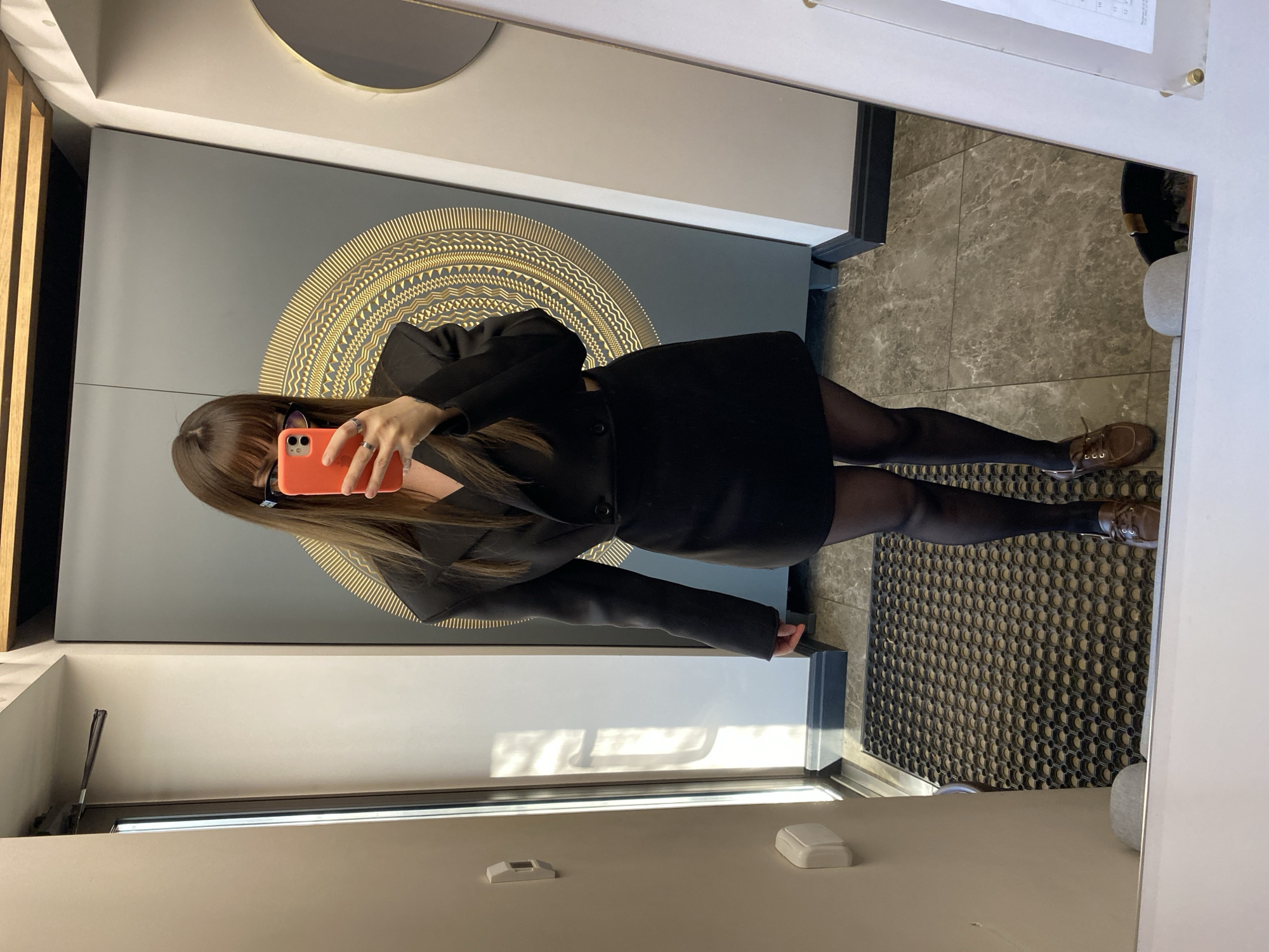 A woman is taking a selfie in a mirror. The mirror's reflection shows a gray wall with a circular gold design. The woman is wearing a black blazer and skirt with black tights and brown shoes. The location cannot be determined as Votkinsk, Russia.