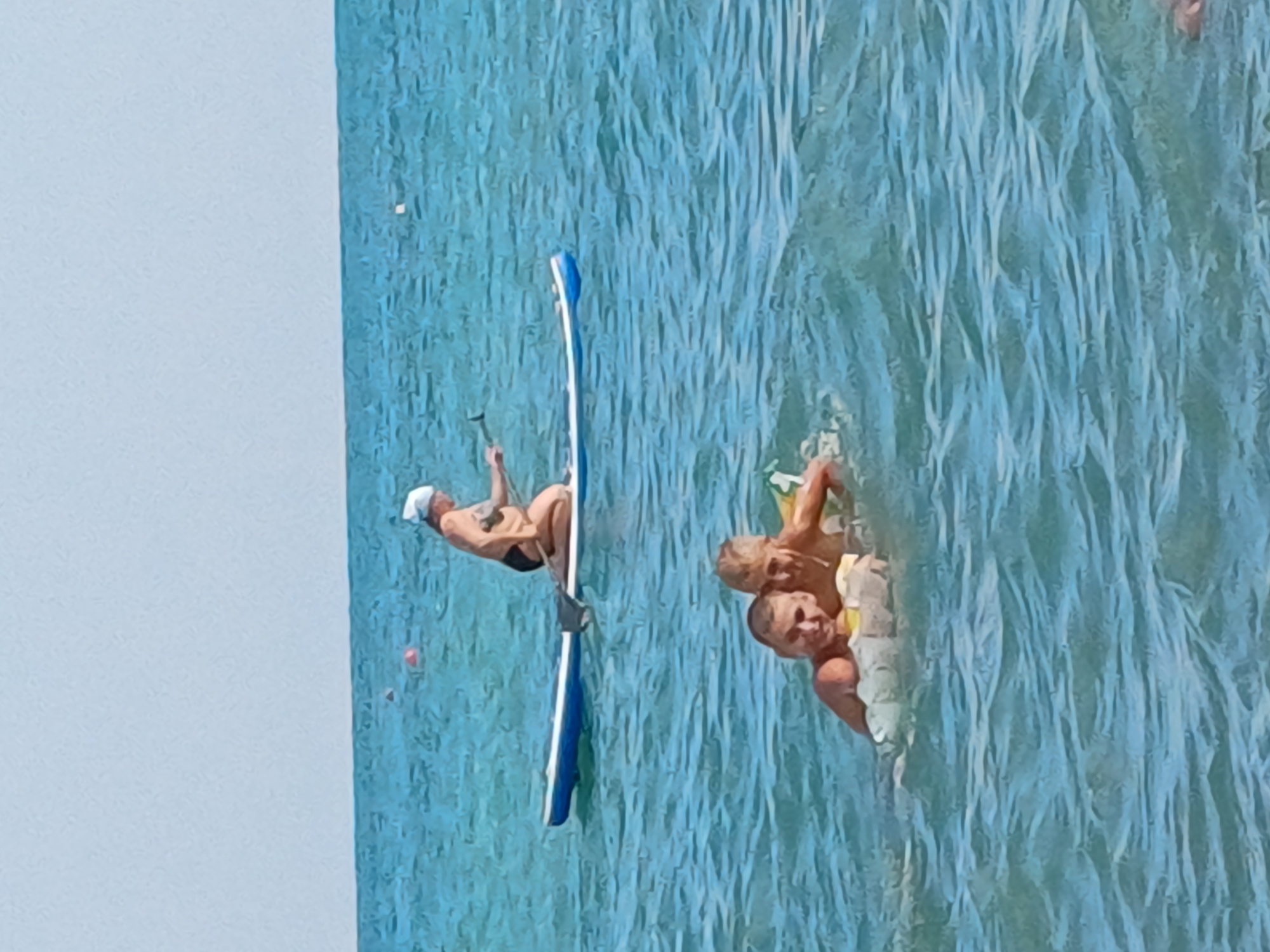 A woman wearing a white headscarf and dark swimsuit is kneeling on a paddleboard in the turquoise ocean. She is holding a paddle and appears to be propelling herself through the water. Two children are floating in the water in the foreground, appearing to be in a small inflatable device. One child is looking at the camera, while the other is partially obscured. The scene takes place on a clear day, with a calm ocean and a pale blue sky. This scene is likely taking place on the coast of Sochi, Russia, known for its beaches and water activities.