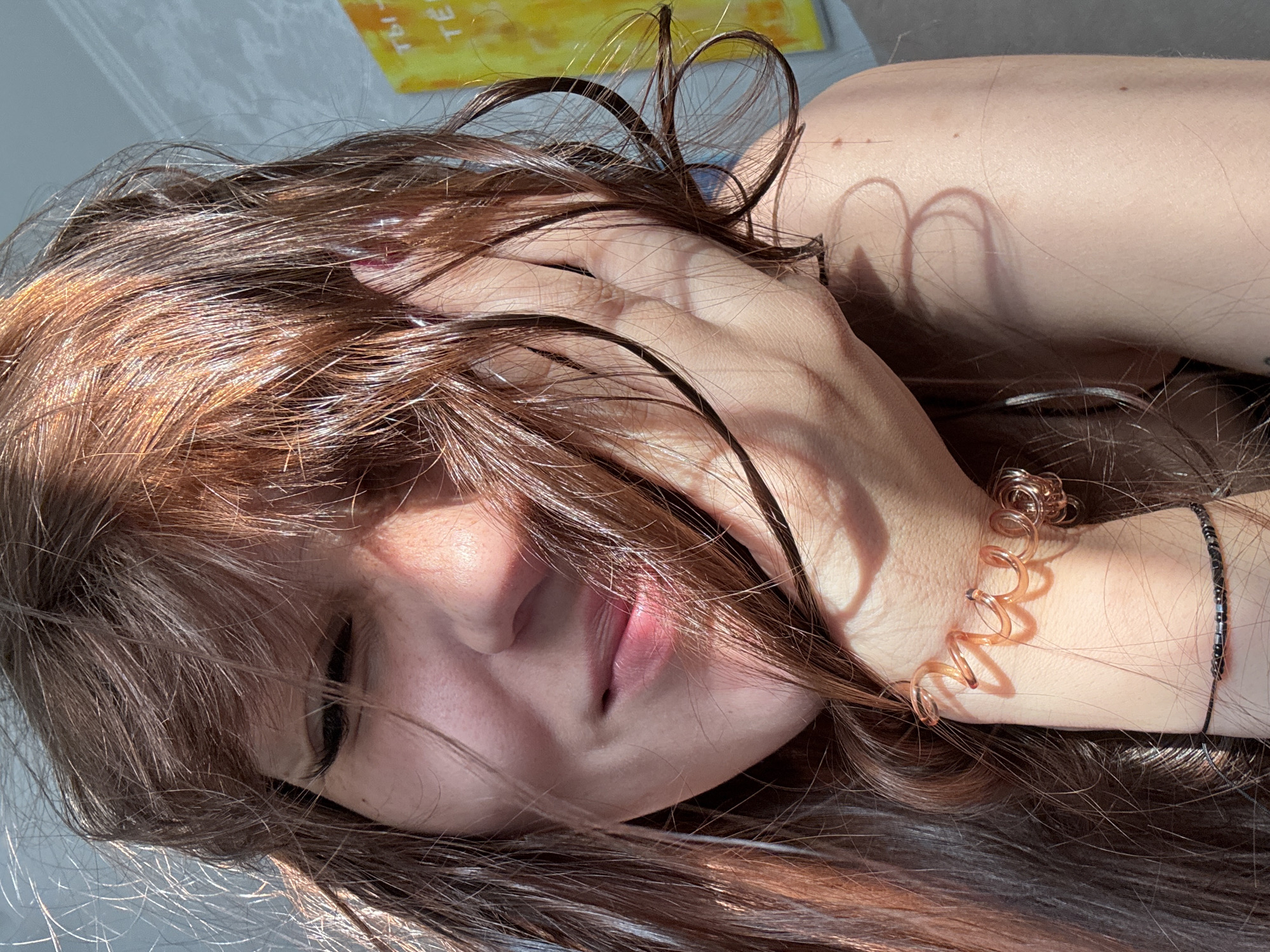 This is a close-up, vertical, eye-level shot of a young woman lying down, with her head turned to her right, as if resting. She has long, dark brown hair that is slightly damp and falls around her face. Her eyes are closed, and her lips are slightly parted, suggesting a relaxed or pensive state. Sunlight casts shadows on her face and arm, creating a warm and intimate atmosphere. One shadow, formed by her hair, creates a heart shape on her arm. She is wearing a copper-colored spiral bracelet on her left wrist, and a thin black bracelet with small beads on the same wrist. The background appears to be a light-colored surface, possibly a bed or a table, with a hint of a yellow object with some text on it in the upper background. The lighting indicates it is daytime, likely late morning or early afternoon, with bright sunlight. The mood is serene and peaceful. There is some visible text on a yellow object in the background, but it is blurry and difficult to decipher.