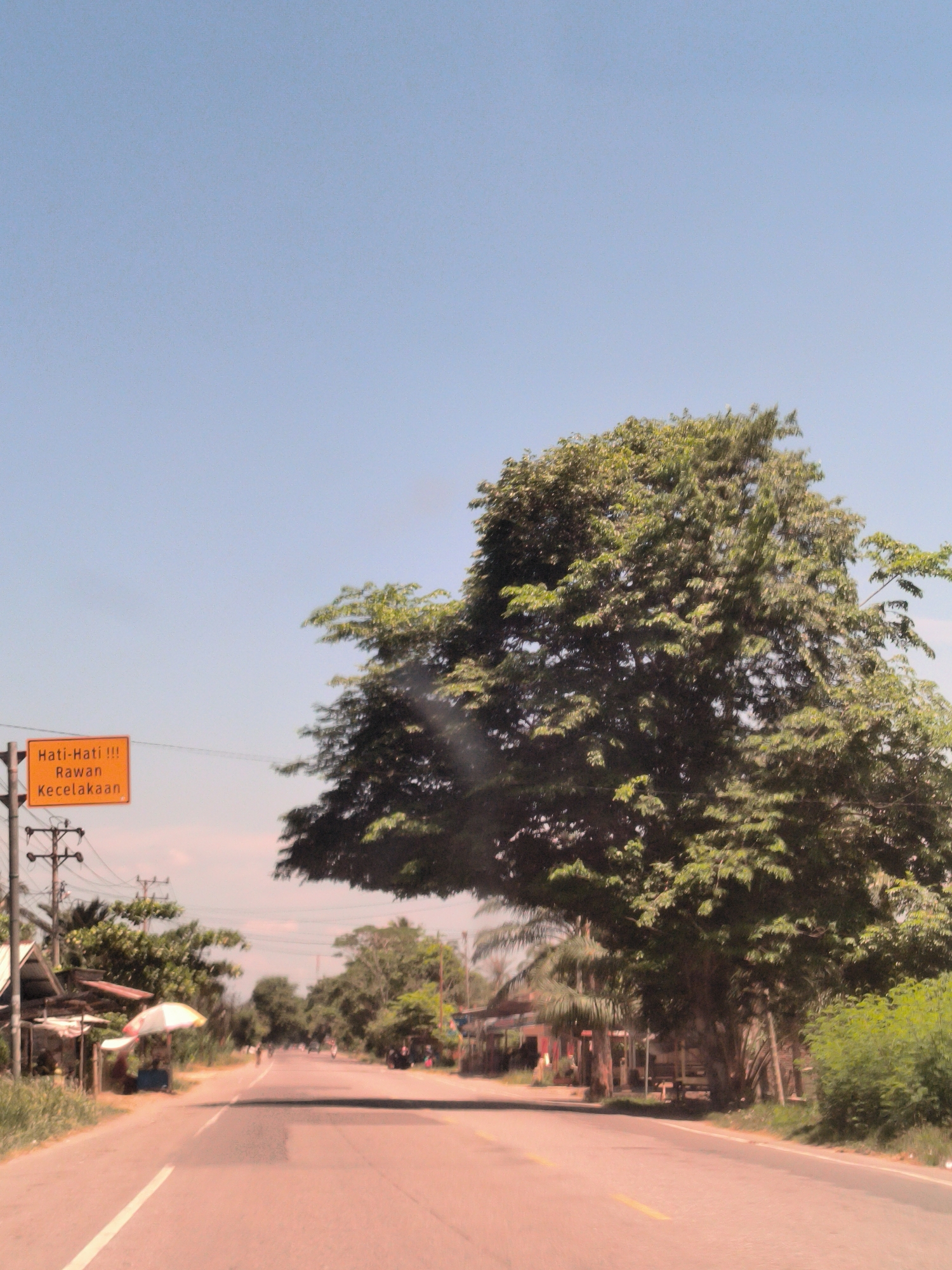 The image shows a road in Matangpanyang, Indonesia. The road is asphalt, with a yellow centerline. On the left side of the road is a yellow sign with black text that says "Hati-Hati !!! Rawan Kecelakaan", and there are power lines and some small buildings. On the right side of the road is a large green tree. The sky is blue and clear. The scene suggests a rural or suburban area.