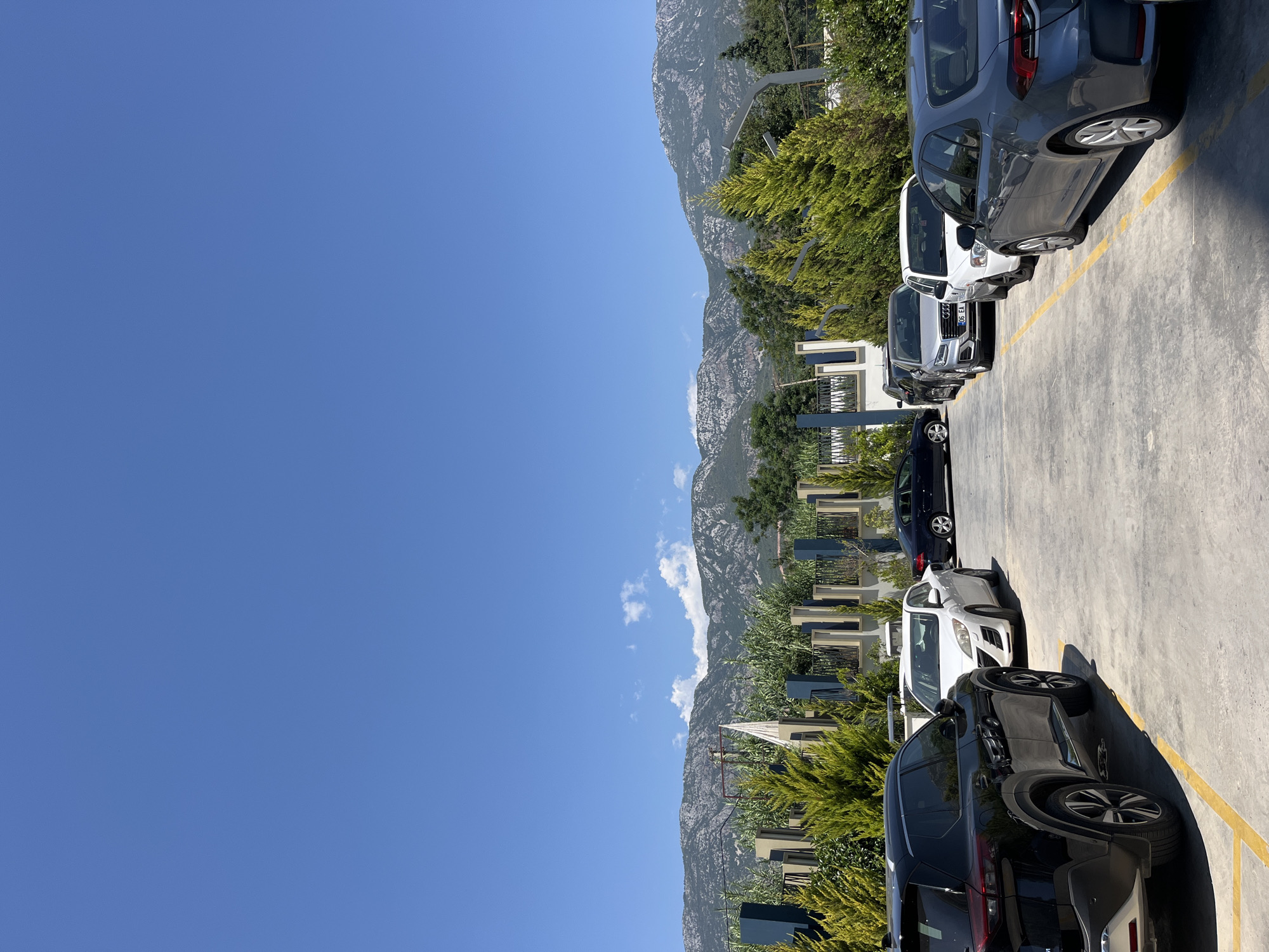 The image shows a parking lot with several cars parked in a row under a clear blue sky. In the background, there are mountains with some vegetation and trees. There are also some modern buildings with abstract architectural elements and fences. The cars are of various colors and models, including a black SUV, a white sedan, a silver sedan, and a dark grey sedan. The parking lot appears to be paved with concrete and marked with yellow lines. The scene suggests an outdoor environment, possibly in a residential or commercial area of Alanya, Turkey, given the presence of mountains and the architectural style. No people are visible in the image.