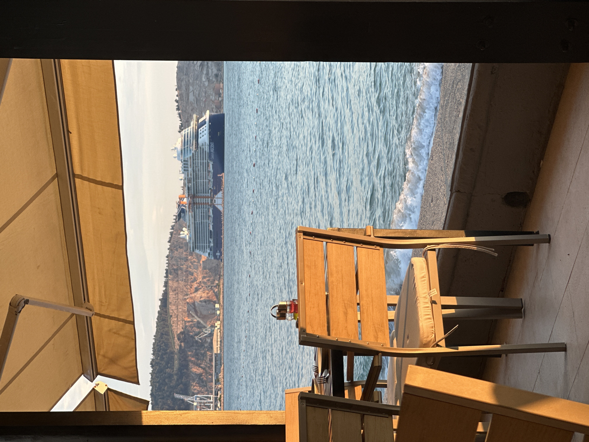 This photo was taken from a covered outdoor dining area in Šušanj, Montenegro. The dining area has light-colored wood and metal patio furniture. A wooden-slatted table and two matching chairs are in the foreground. At the table, there is a glass bottle with a red cap. The photo appears to have been taken at sunset.
In the distance, over the water, are land formations and a large ocean liner. The water is choppy, with white waves. There are white barriers to the right of the image, which seem to follow the line of the sea.