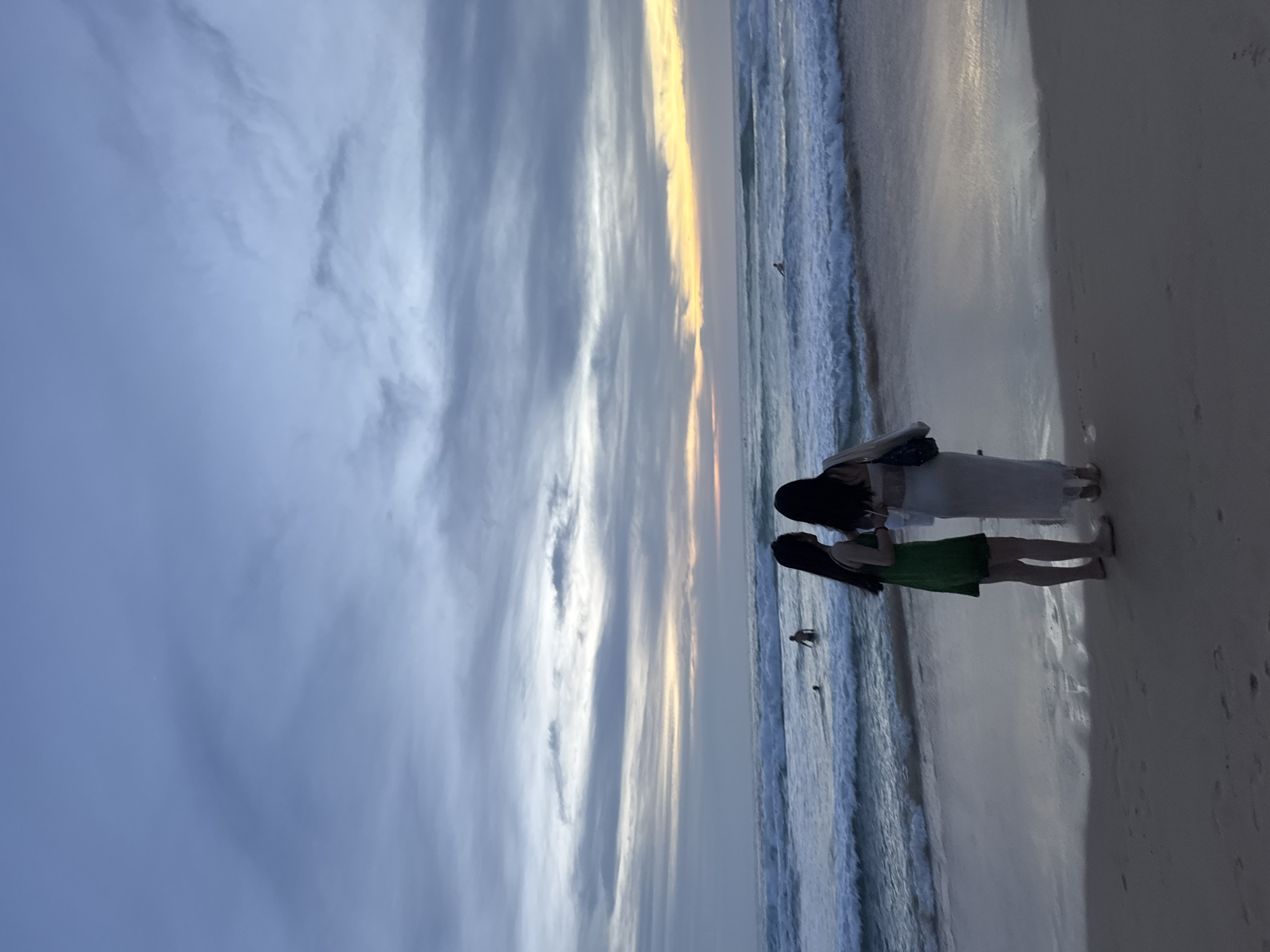 The image shows a beach scene at dusk in Karon, Thailand. Two women are standing on the sand, facing the ocean. The woman on the left is wearing a green dress, and the woman on the right is wearing a white dress. Both women have long dark hair. The ocean has moderate waves, and several people are in the water, some appearing to be swimming or wading. The sky is filled with dramatic clouds, with a sliver of orange and yellow light visible near the horizon, indicating the setting sun. The sand in the foreground is wet and reflects the sky and ocean.