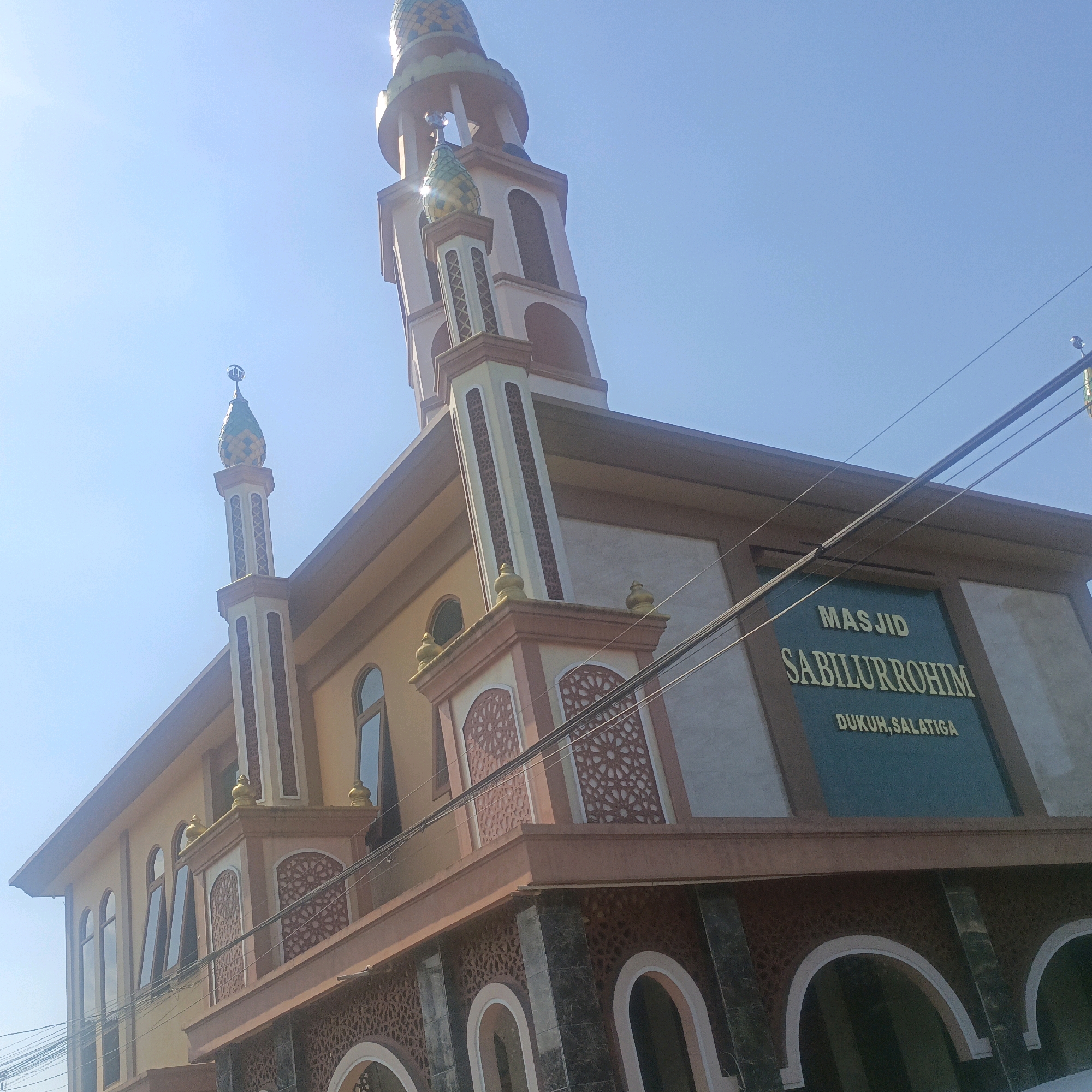 The image shows the exterior of a mosque named Masjid Sabilurrohim, located in Dukuh, Salatiga, Indonesia. The mosque features a tall minaret with a decorative pointed roof, and a larger, more prominent minaret also with a decorative pointed roof. The building itself is multi-colored, with a tan main body and brown and pink accents. Intricate decorative patterns are visible on various parts of the building, including above arched doorways and on the sides of the minarets. The building has several arched windows. A sign on the building reads "MASJID SABILURROHIM DUKUH, SALATIGA". The sky is clear and blue, suggesting a sunny day. There are power lines visible in the foreground. No people are present in the image.