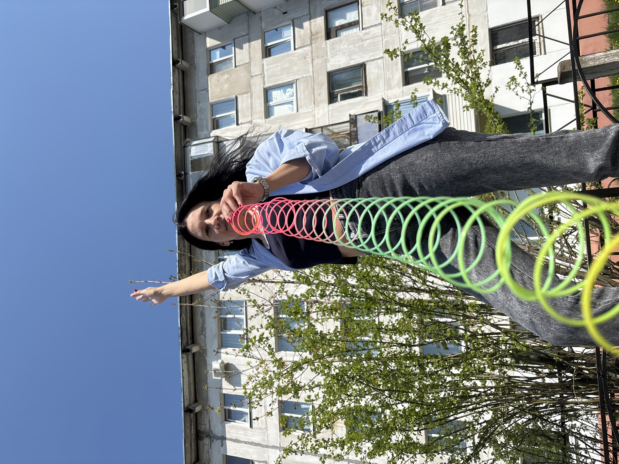 A woman stands in front of an apartment building, holding a colorful slinky toy. She is wearing a blue button-down shirt, a black top, and gray jeans. Her dark hair flows around her face. The apartment building behind her has multiple windows. There is a small tree with green leaves to the right of the building. The sky above is a clear blue. The location appears to be in an urban area. The photo includes a wrought iron fence or railing in the lower right corner. The image orientation suggests the photo was taken from a lower angle. It is daytime. Based on the style of architecture, this could be Chaikovsky, Russia.