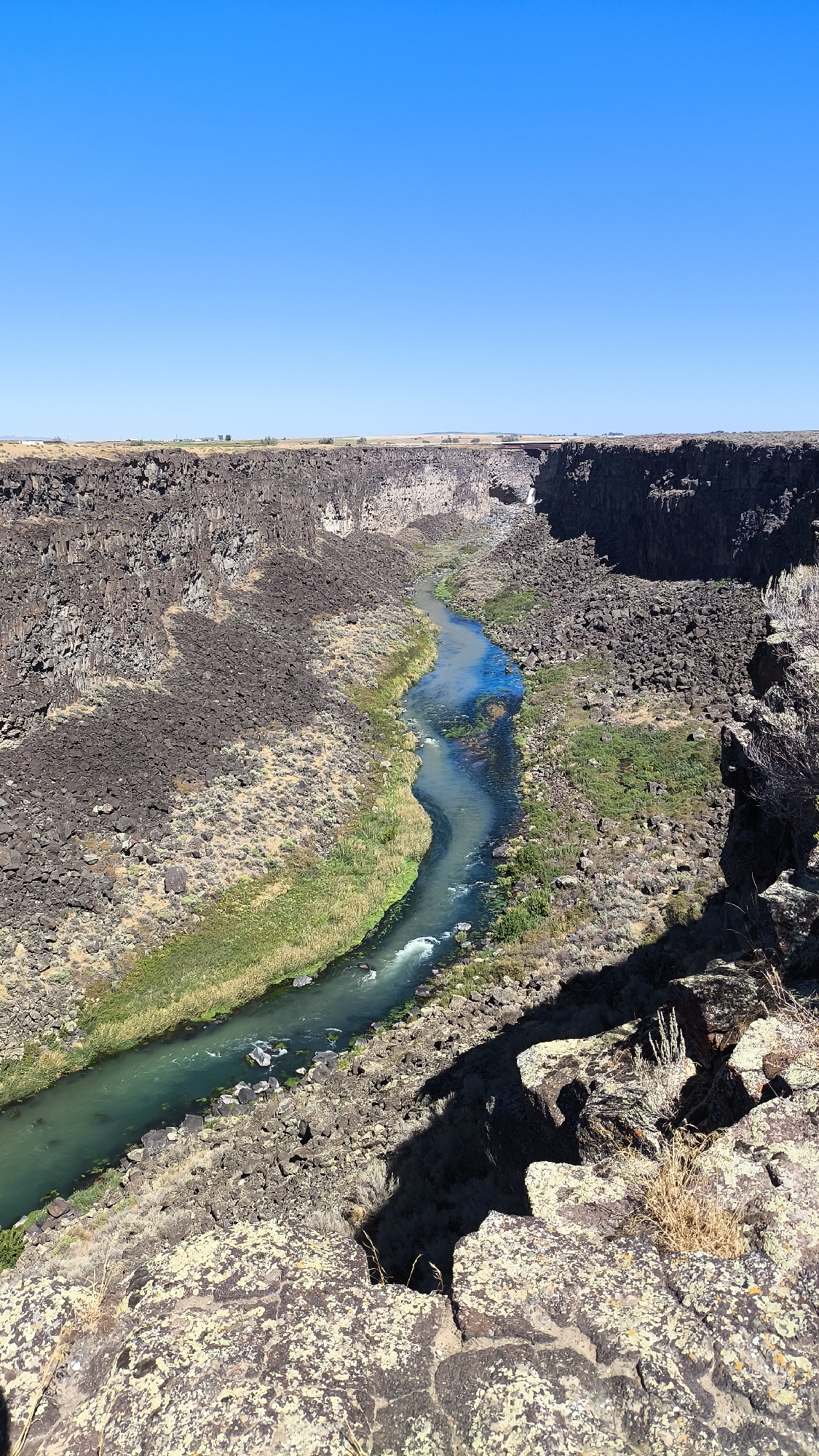 An aerial view captures a winding, turquoise river flowing through a deep canyon under a clear blue sky. The canyon walls are rugged and composed of dark, volcanic rock, with patches of dry, sparse vegetation and light brown soil clinging to the slopes. Green reeds and grasses line the riverbanks, creating a contrast with the barren rock. The river itself is dynamic, with visible ripples and white water indicating flow. In the distance, the canyon opens up to a flat, arid landscape with a few scattered trees and buildings, suggesting it is on the outskirts of a rural area. The foreground consists of rough, textured rock formations, casting long shadows from the bright sunlight. The scene appears to be a natural landscape, likely a state park or natural monument, given its grandeur and the preservation of its features. Based on the prompt specifying Tuttle, United States, this scene depicts a natural landmark within or near that city.