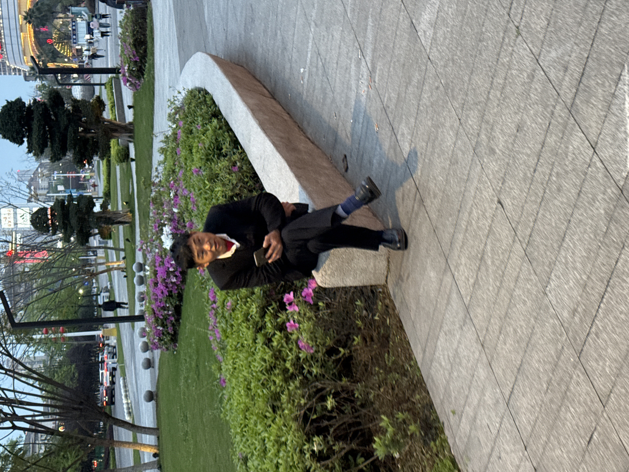 An Asian man wearing a black suit, black shoes, and blue socks is sitting on a concrete ledge in a park-like setting. He is holding a mobile phone in his left hand. The ledge is situated next to a flower bed with green bushes and purple flowers. Beyond the flower bed is a grassy area with trees and street lights. In the background, cars are visible on a road. In the far background, high-rise buildings are present with lights illuminating them. The location appears to be in an urban area within the city of Jiangjin, China, based on the architecture and environment. The time of day is likely late afternoon or early evening.