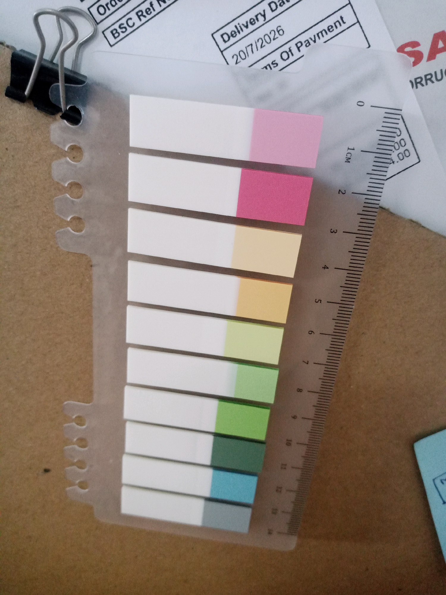 A close-up, top-down shot captures a small binder clip holding together documents and a set of colorful sticky notes. The documents, partially visible, have printed text and dates, one reading "BSC Ref N" and another "Delivery Dat 20/7/2026". Adjacent to the sticky notes is a clear ruler marked with centimeters, indicating measurements from 0 to 14. The sticky notes themselves are arranged in a vertical stack, with each note featuring a white portion followed by a colored band, including pink, light orange, light green, and blue. The background appears to be a cardboard surface. The scene does not provide any information about the city of Balaraja, Indonesia.