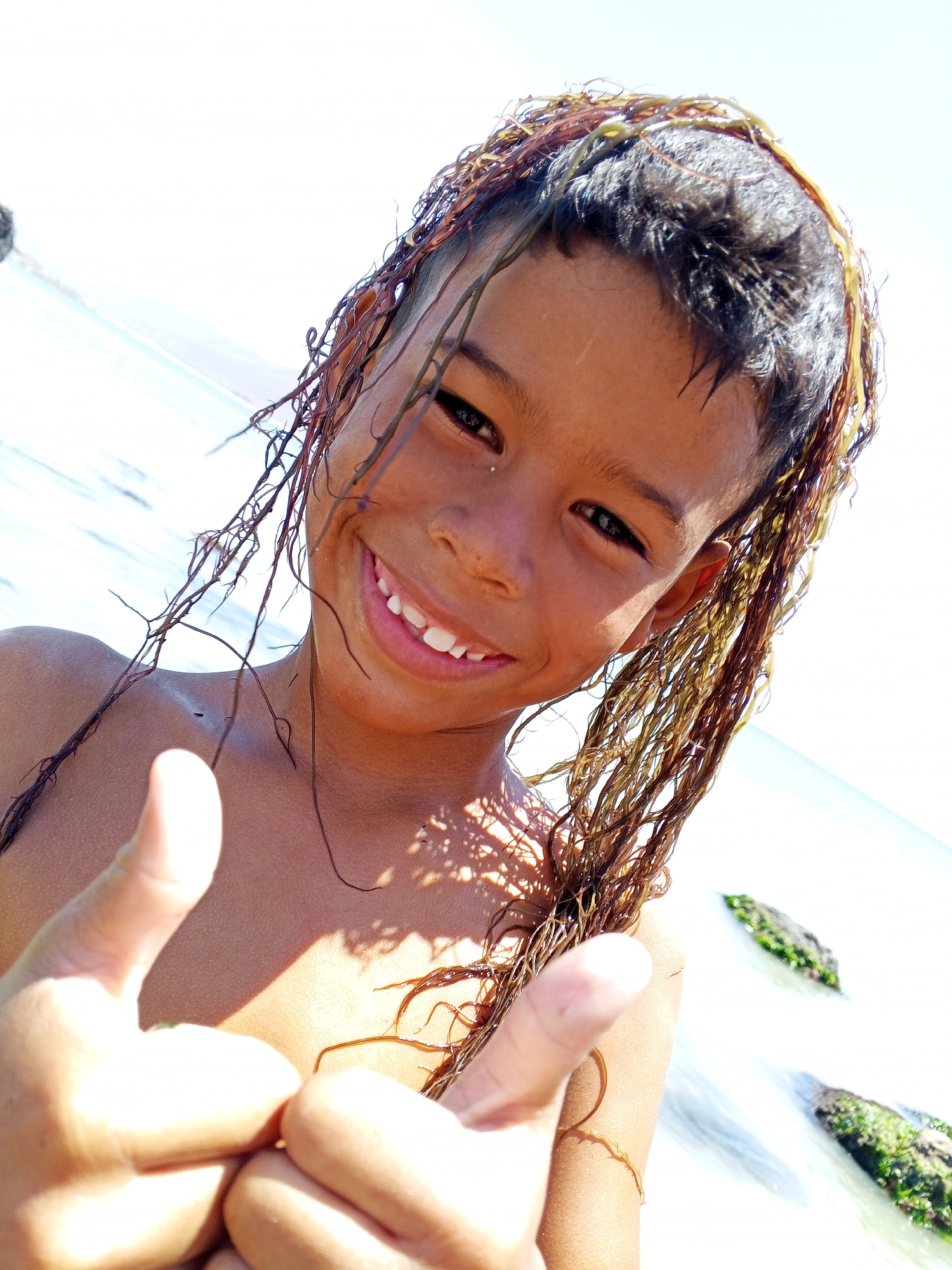 A young person with dark hair and seaweed draped over their head smiles at the camera and gives a thumbs-up gesture. The background appears to be a beach with rocks visible in the water. The location is Unknown, Unknown.
