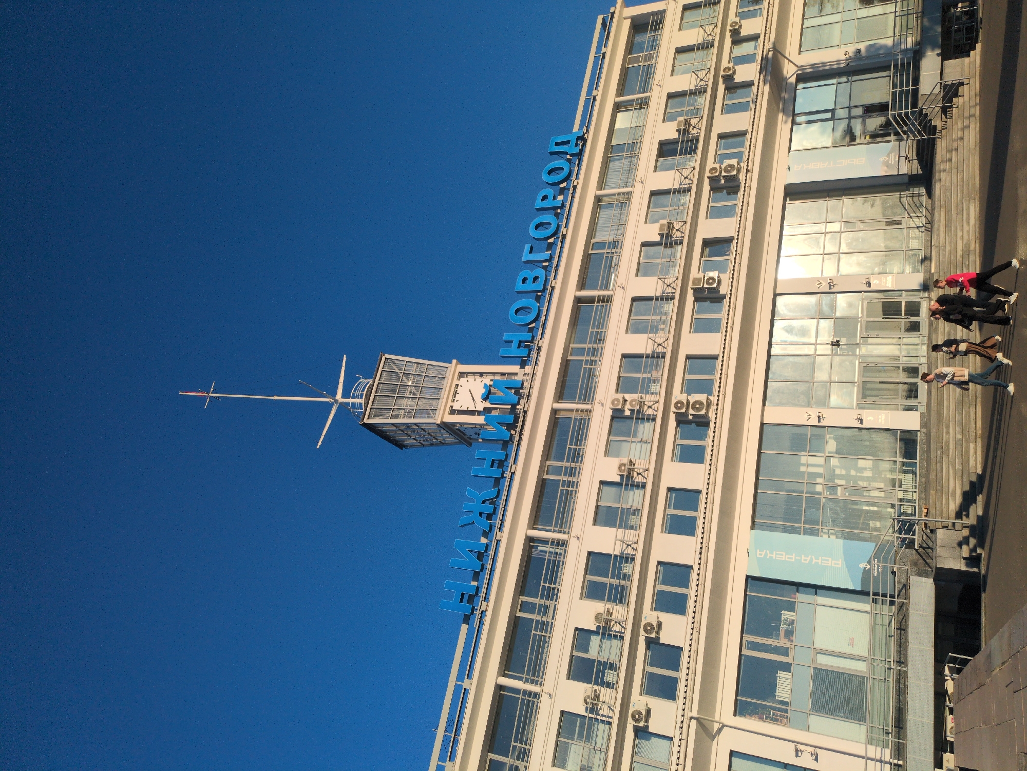 The image captures the Nizhny Novgorod river terminal building against a clear blue sky. The building is a multi-story structure with a white facade and numerous windows, some of which are air-conditioned units installed externally. Prominently displayed on the top of the building are large blue letters spelling out "НИЖНИЙ НОВГОРОД" (Nizhny Novgorod). A tower-like structure with a mast stands on the roof of the building. In front of the building, several people are walking on a paved area and a set of stairs. One banner with Cyrillic text, "РЕКА-РЕКА," is visible on the glass facade of the building. The overall scene suggests a sunny day at a significant architectural landmark in Nizhny Novgorod, Russia.
