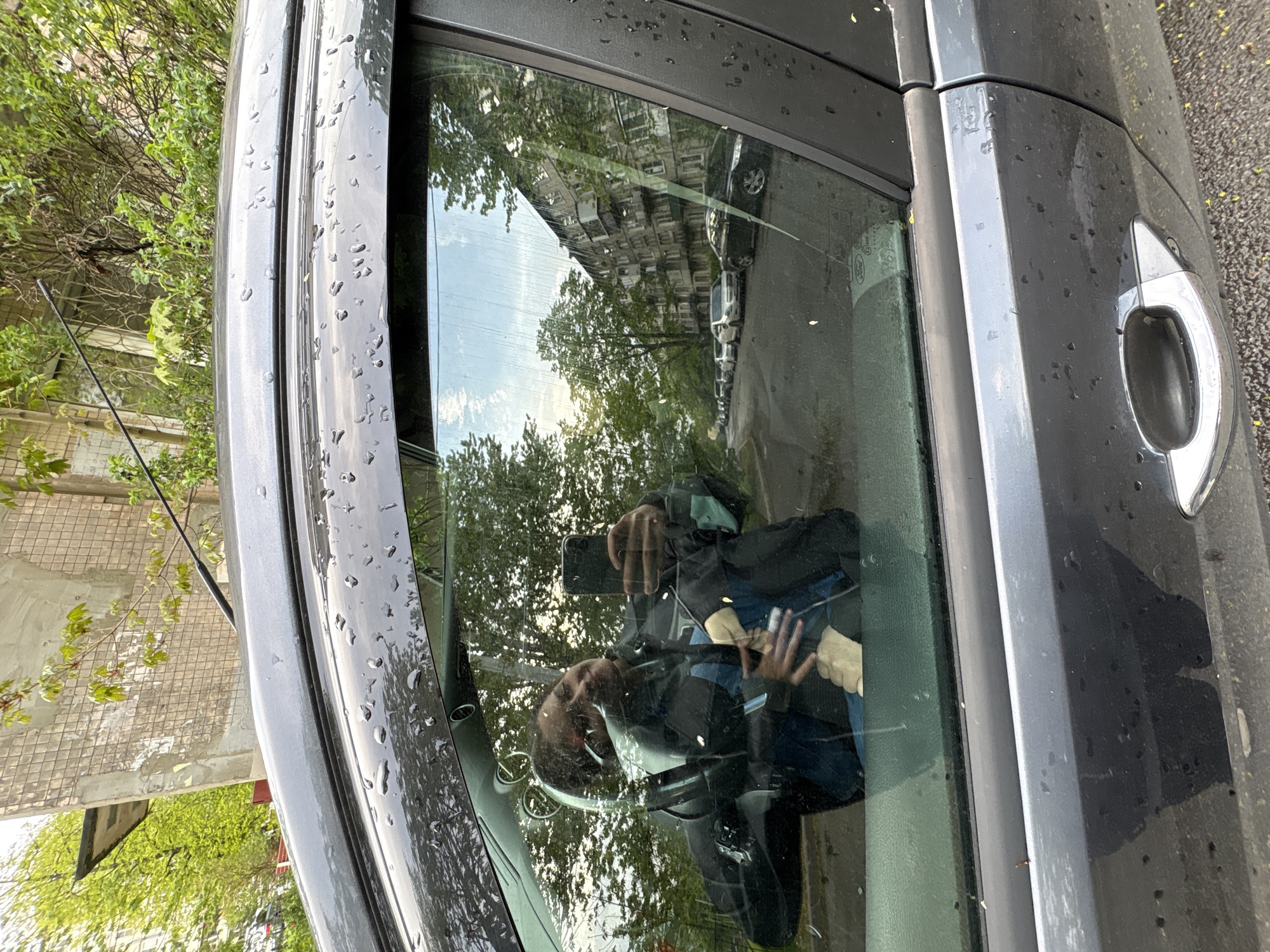 The image shows the exterior of a dark-colored car, likely parked on a street in Saint Petersburg, Russia, judging by the architecture reflected in the car window. The reflections in the window include buildings, trees, and a row of parked cars. Raindrops are visible on the car's surface. A person is reflected in the window, holding a smartphone and taking the picture. The scene appears to be an urban environment.