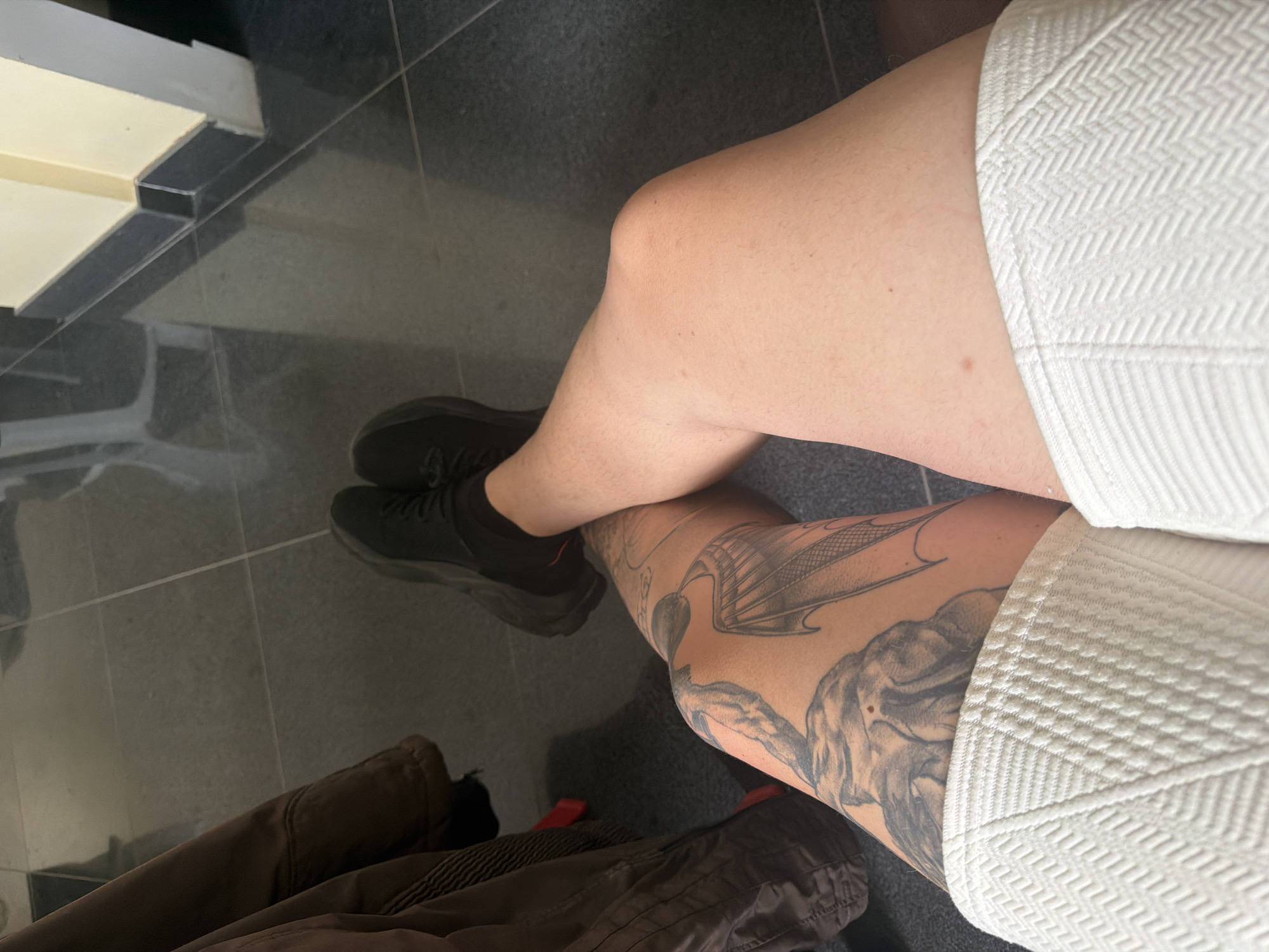 The image depicts a person's legs from the knee down, seated on a floor with dark tiled squares. The person is wearing black athletic shoes and white textured shorts. The left leg has a large tattoo on the thigh featuring what appears to be a dragon's wing and possibly an elephant's head. The right leg is bare and smooth. A brown jacket or coat is visible on the left side of the frame. The background shows a wall with a yellow upper section and a dark baseboard, with the floor extending into the distance.