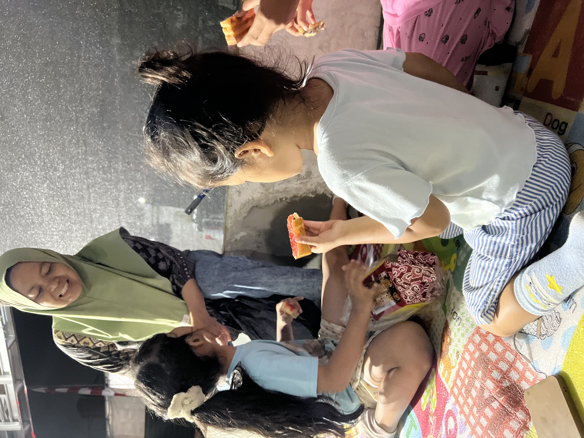 In Klapanunggal, Indonesia, an outdoor scene depicts three individuals, likely a family, gathered on a colorful mat. A woman wearing a light green hijab and a patterned top is interacting with two young girls. One girl, with long dark hair tied in a ponytail adorned with a white bow, wears a light blue t-shirt and patterned shorts. The other girl, facing away from the camera, has her dark hair in a bun and is wearing a white t-shirt and striped blue shorts. Both girls are holding and eating pieces of cake. The environment appears to be an open area, possibly a yard or patio, with a textured gray wall in the background and some fencing visible. The lighting suggests it is either dusk or evening.