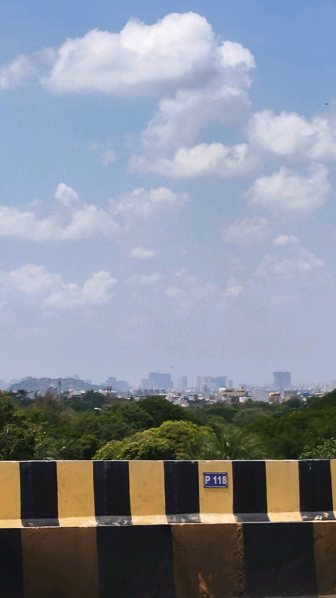 The image captures a hazy view of the Hyderabad skyline under a bright blue sky with scattered white clouds. In the foreground, a concrete barrier painted with alternating yellow and black stripes is visible, with a small blue sign bearing the white text "P 118" attached to its upper section. Beyond the barrier lies a dense expanse of green trees and foliage. In the distance, the city of Hyderabad presents a silhouette of buildings, with taller structures, possibly skyscrapers, piercing the hazy atmosphere. The overall impression is that of a typical cityscape on a clear but hazy day, viewed from an elevated position.