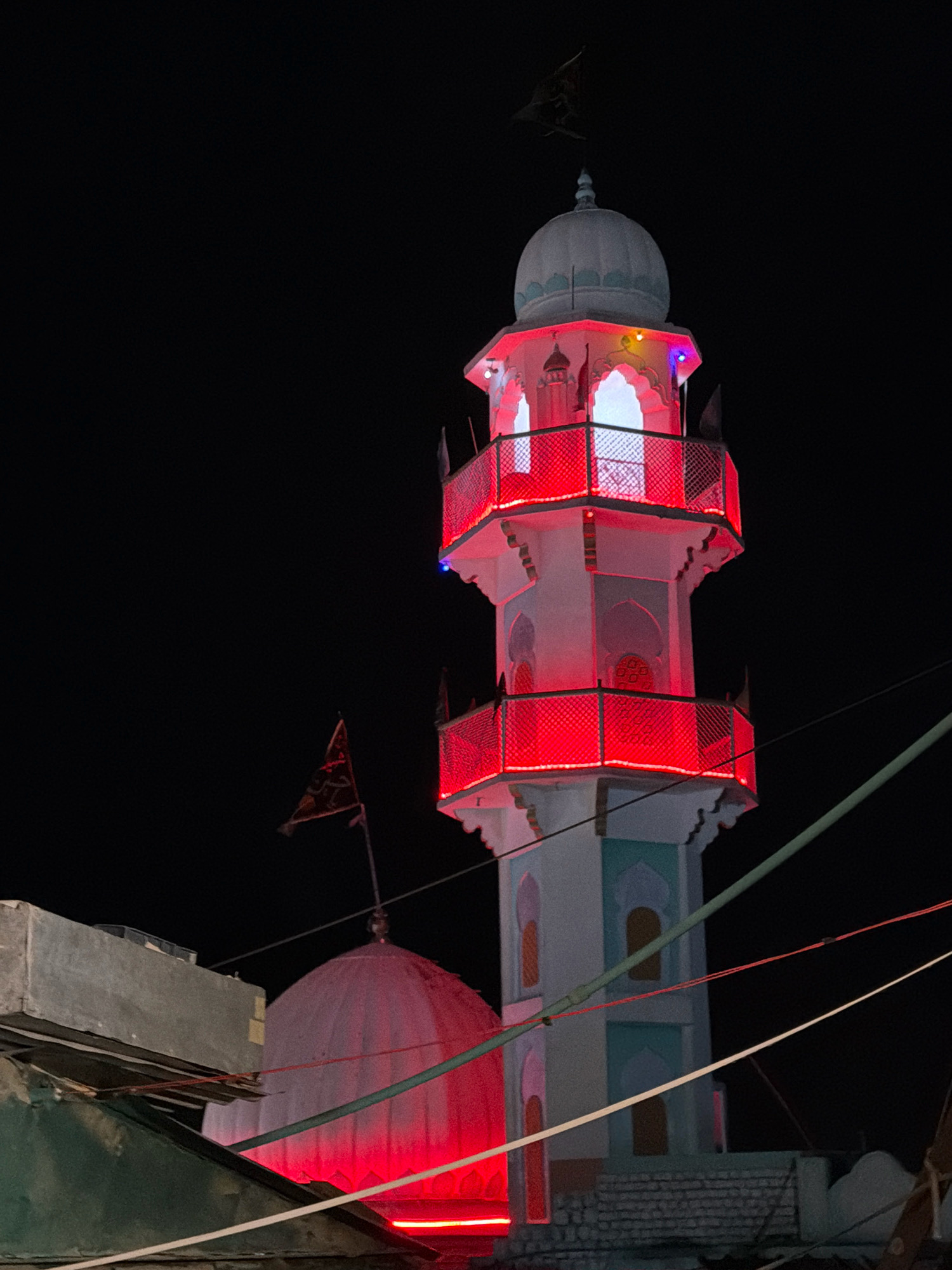 The image shows a brightly lit minaret of a mosque at night, located in Leh, India. The minaret is white with red lights outlining its balconies and arched windows, and it is topped with a pale blue dome. A flag with intricate designs is flying from the top of the minaret. To the left of the minaret is a red-lit dome of another structure, possibly the main mosque building, and a gray concrete structure. The scene is dark, indicating it's nighttime, with visible power lines crisscrossing the frame. There are no people visible in the image.