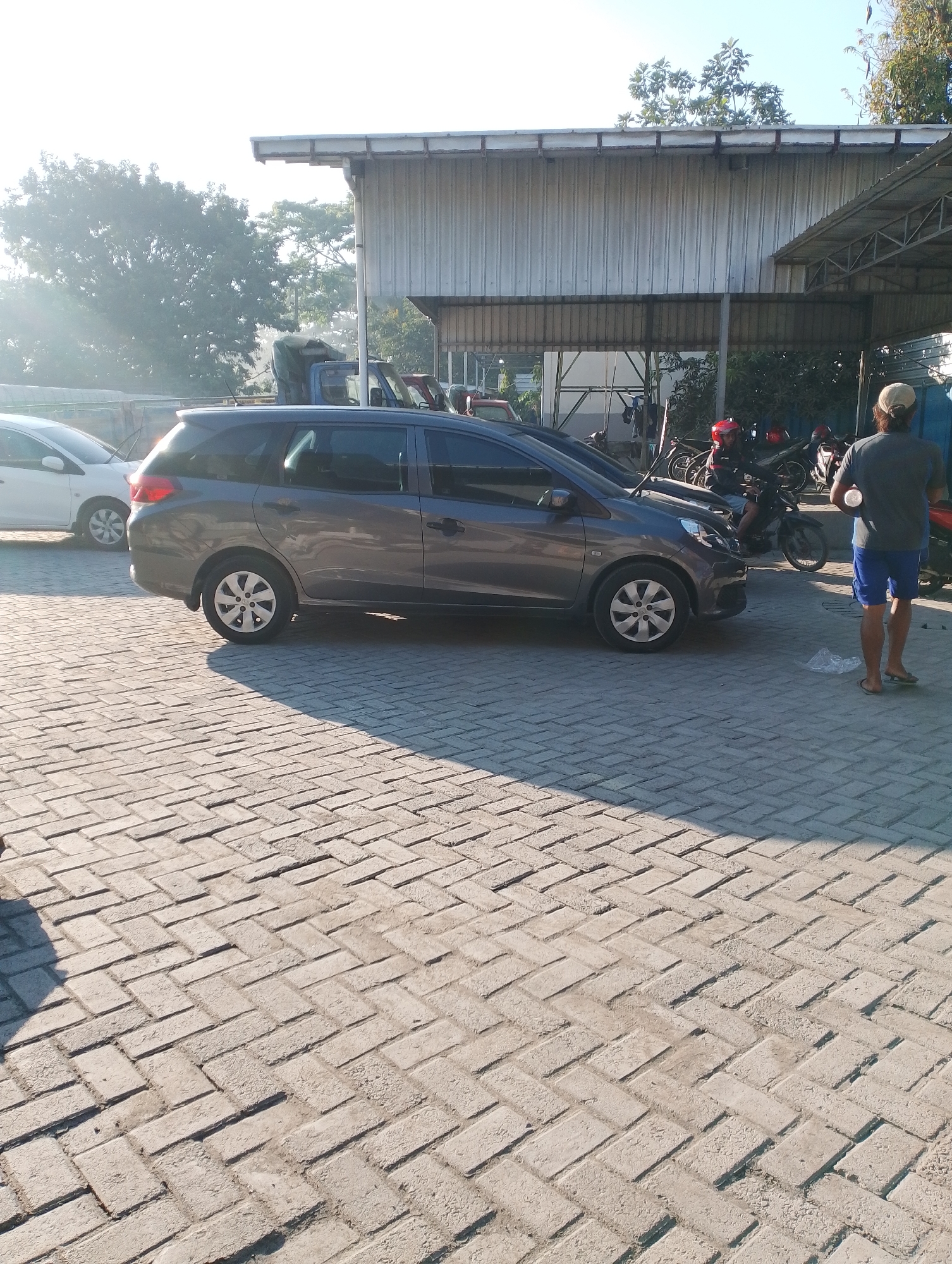 In Jambean, Indonesia, a grey Honda Mobilio is parked on a herringbone-patterned brick paver surface in front of a corrugated metal building with a partial awning. A person wearing a light-colored cap, grey t-shirt, and blue shorts stands to the right of the Mobilio, facing away from the camera and holding a white object. Several motorcycles are visible parked in front of the building, one with a rider wearing a red helmet. The scene is illuminated by natural sunlight, casting shadows on the ground, and the sky is clear.