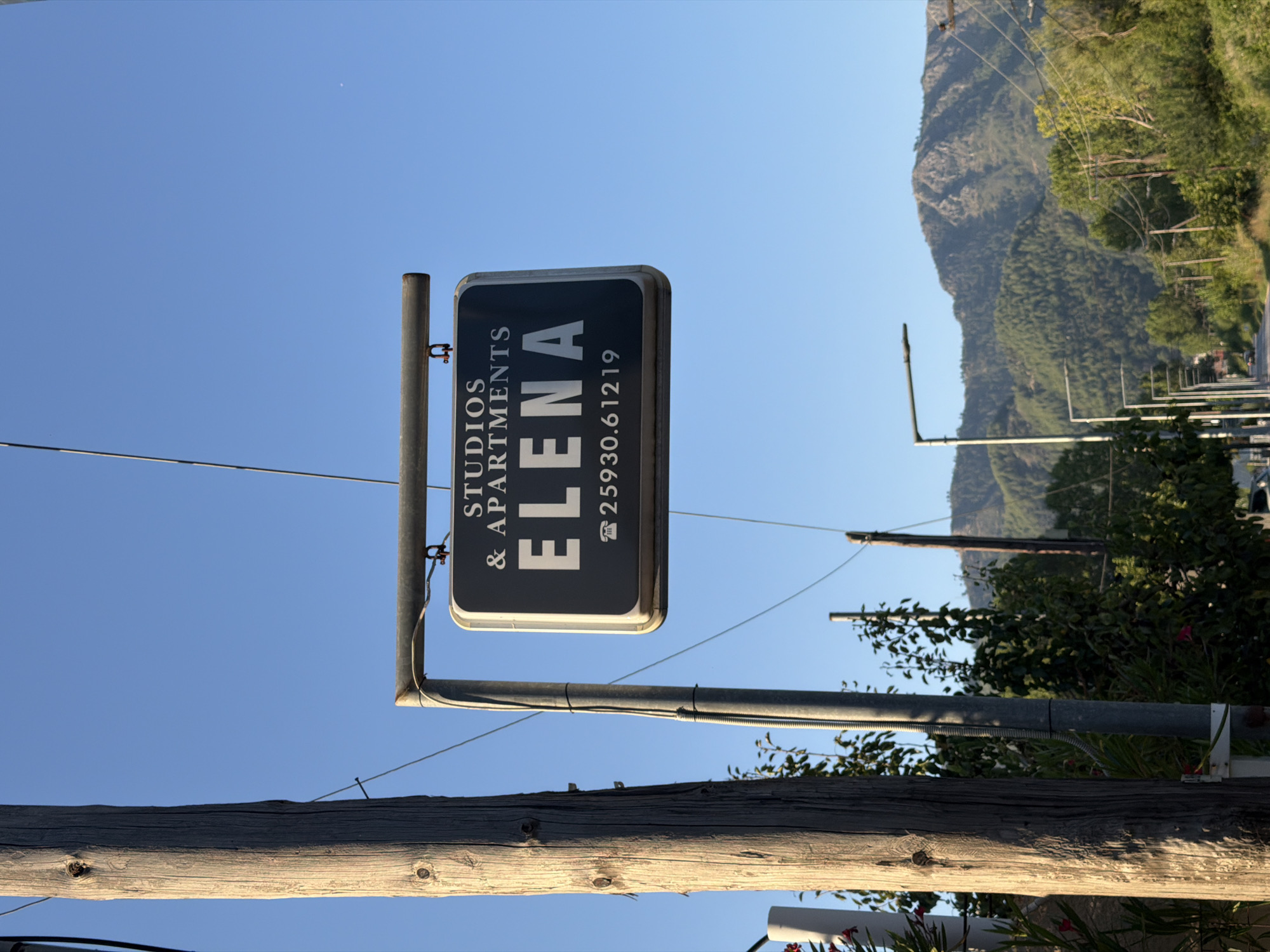The image shows a sign for "ELENA STUDIOS & APARTMENTS" in Golden Beach, Greece. The sign is rectangular with a dark background and white lettering. The words "STUDIOS" and "& APARTMENTS" are at the top, and "ELENA" is in large letters in the center. Below "ELENA" is a telephone icon followed by the number "25930.61219". The sign is suspended from a metal bar attached to a wooden pole and another metal pole. The background is a clear blue sky. In the distance, there are trees and mountains. The scene appears to be along a road.