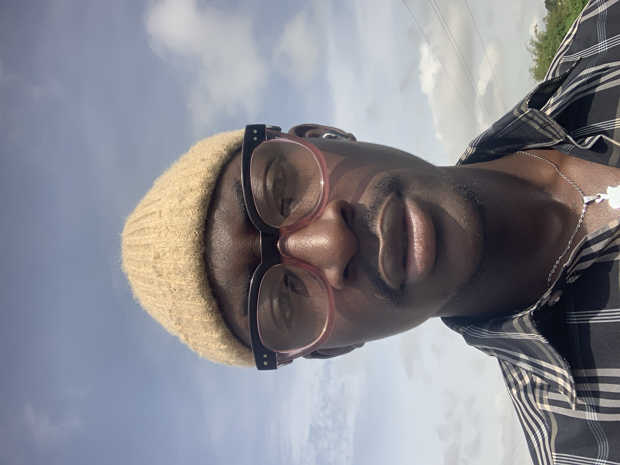 A young dark-skinned man with a light brown beanie, pink-rimmed glasses, and a silver necklace is captured in a close-up selfie. He is wearing a black and white checkered shirt. The background is a blue sky with scattered clouds and some power lines visible. The image appears to have been taken outdoors, possibly in Jos, Nigeria, given the clear skies and the style of the power lines.