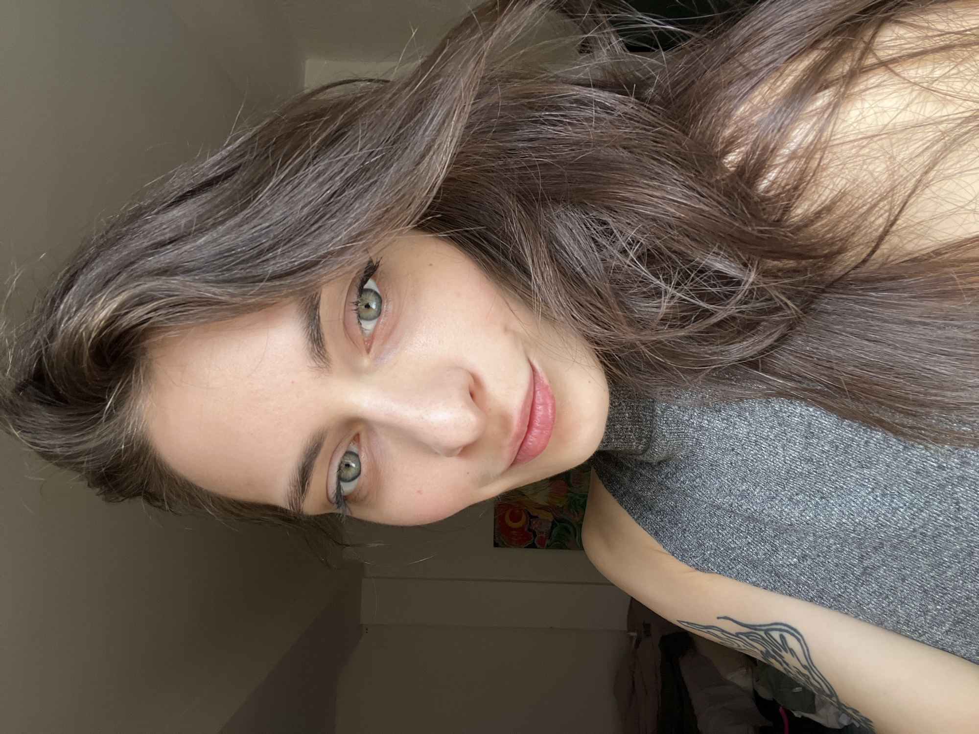 A close-up, angled headshot of a young woman with long, dark brown hair and light green eyes. She is wearing a gray turtleneck sweater. Her skin is fair. She has a tattoo on her left forearm. In the background, there is a wall, part of a painting with red, yellow, and green hues, and a glimpse of a curtain or clothing. It is not possible to determine if the location is in Krasnoyarsk, Russia, based on the visual information provided.