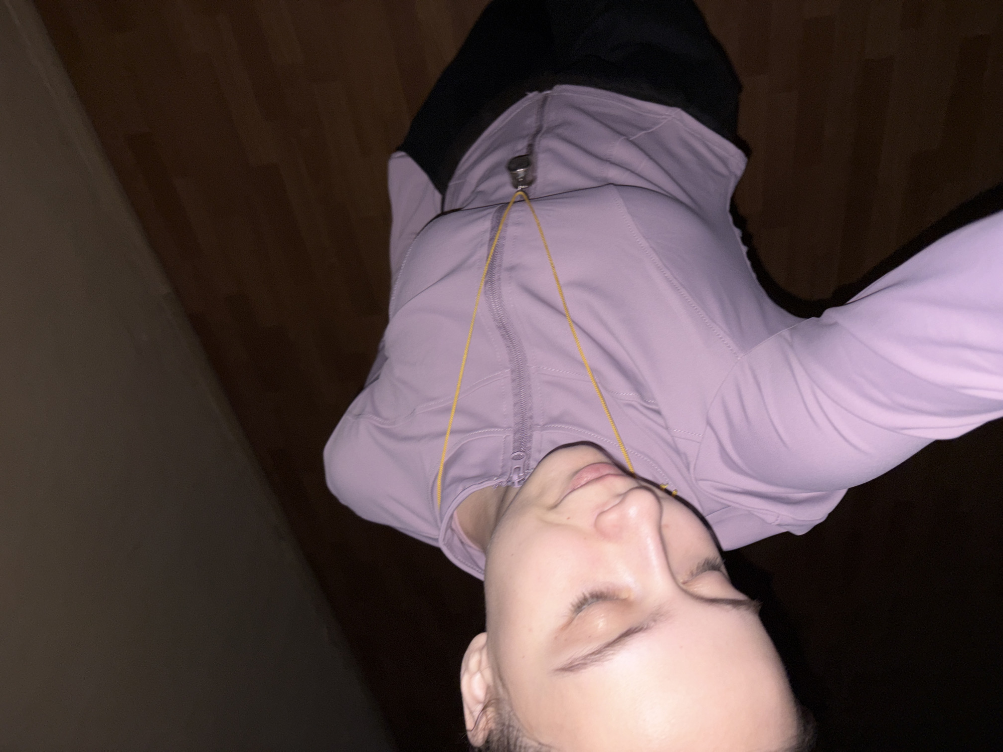 A woman is seen from a high-angle perspective, positioned indoors on a wood floor. She is wearing a light purple zip-up athletic jacket with a yellow drawstring. She appears to be looking down with her eyes closed. The location is Chaikovsky, Russia.