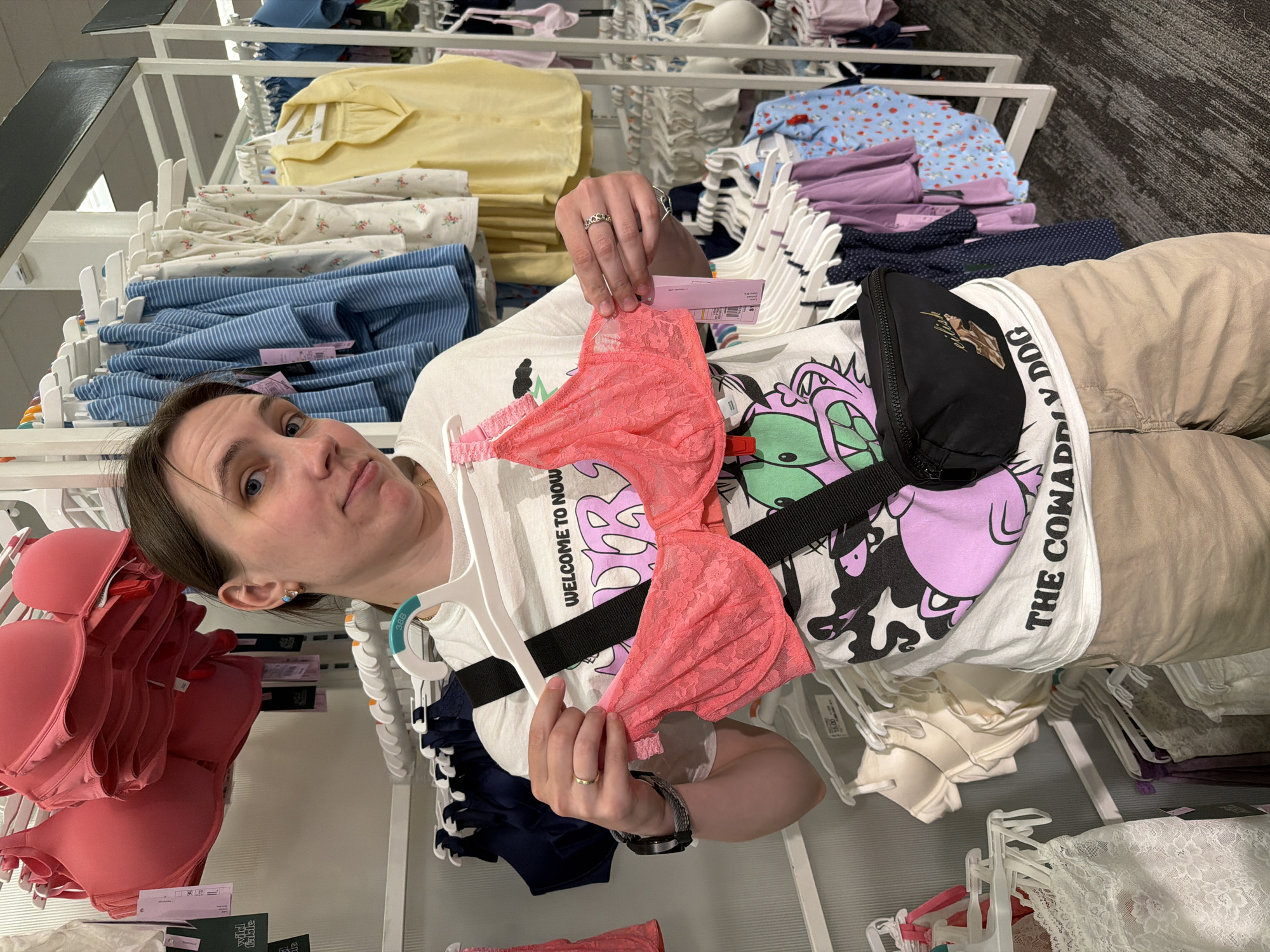A person stands in a retail store, possibly a department store, holding a pink lace bra. The person is wearing a white t-shirt with a graphic print and tan shorts. They have a black fanny pack slung across their chest. Racks of clothing, including bras and other garments, are visible in the background. The environment appears to be an aisle in a store. The image was taken in Beaverton, United States of America.