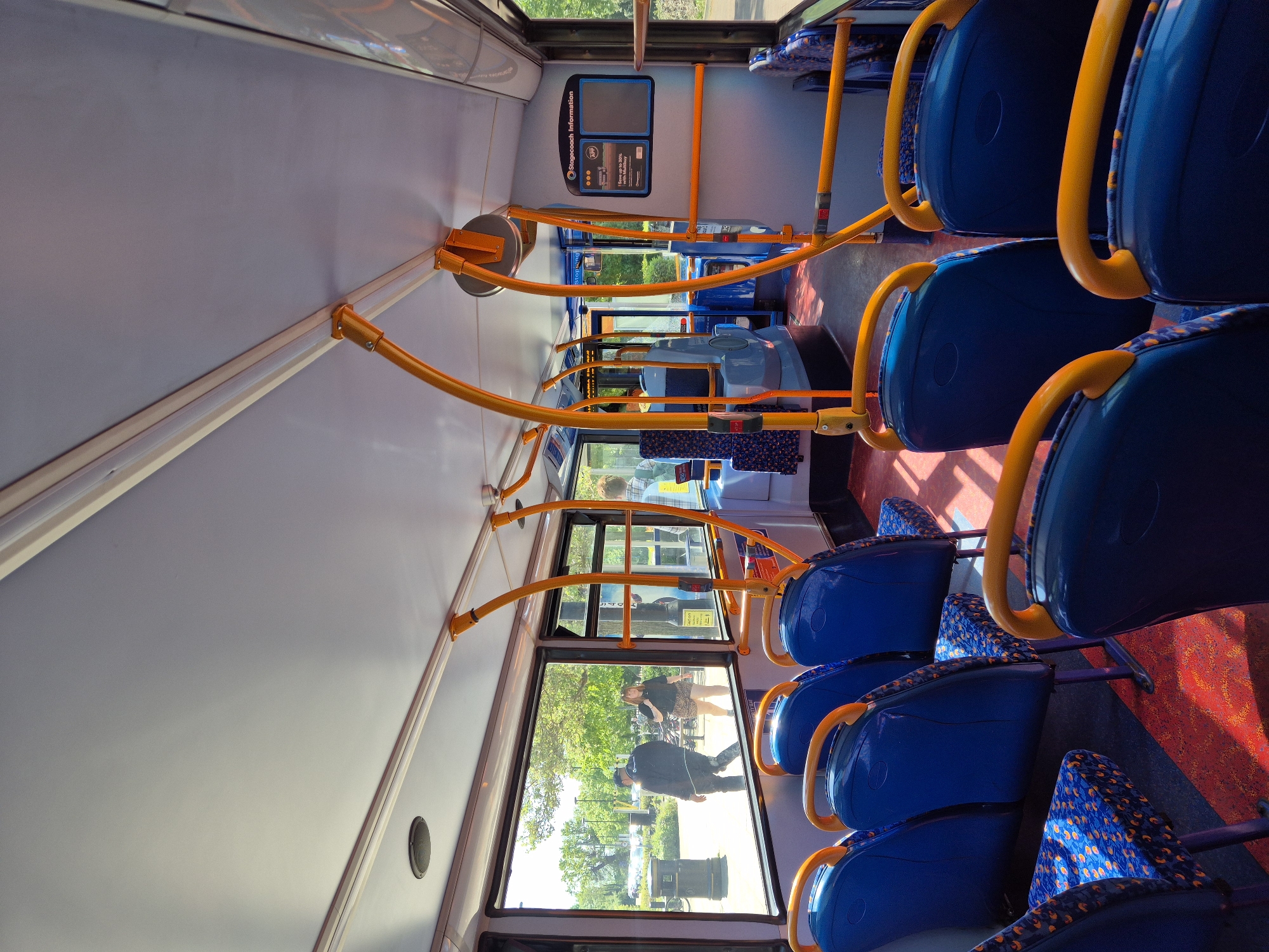 The image shows the interior of a bus, likely a Stagecoach bus, moving through South Cambridgeshire, United Kingdom. Rows of blue seats with patterned headrests line the aisle, with yellow handrails above. Through the bus window, two people are visible outside, one male in a dark jacket and cap walking past a green area and the second person is in a checkered skirt. An information panel is fixed to the wall at the front of the bus. The floor has a patterned red and blue carpet.