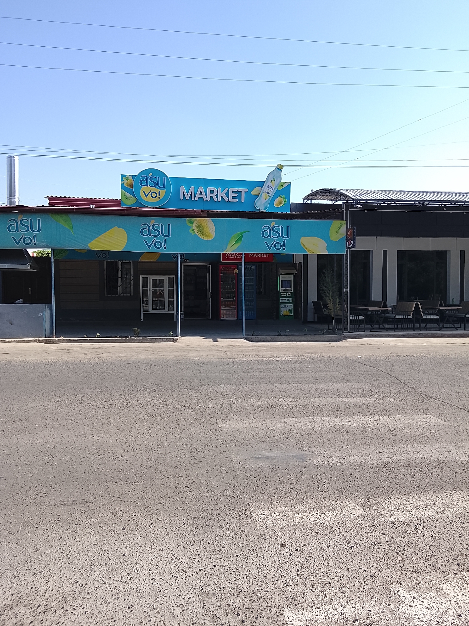 A street-level, eye-level, wide shot captures the exterior of a market in Fergana City, Uzbekistan. The market features a prominent blue awning with the "asu vo! MARKET" logo and fruit imagery. Beneath the awning, the market's entrance is visible, with a Coca-Cola refrigerator to the right and an ATM machine to the left. A traffic light stands in front of the market, near an outdoor seating area with tables and chairs. The street in front of the market is paved with asphalt and shows signs of wear. Overhead, power lines stretch across the clear blue sky. No people are visible in the scene.