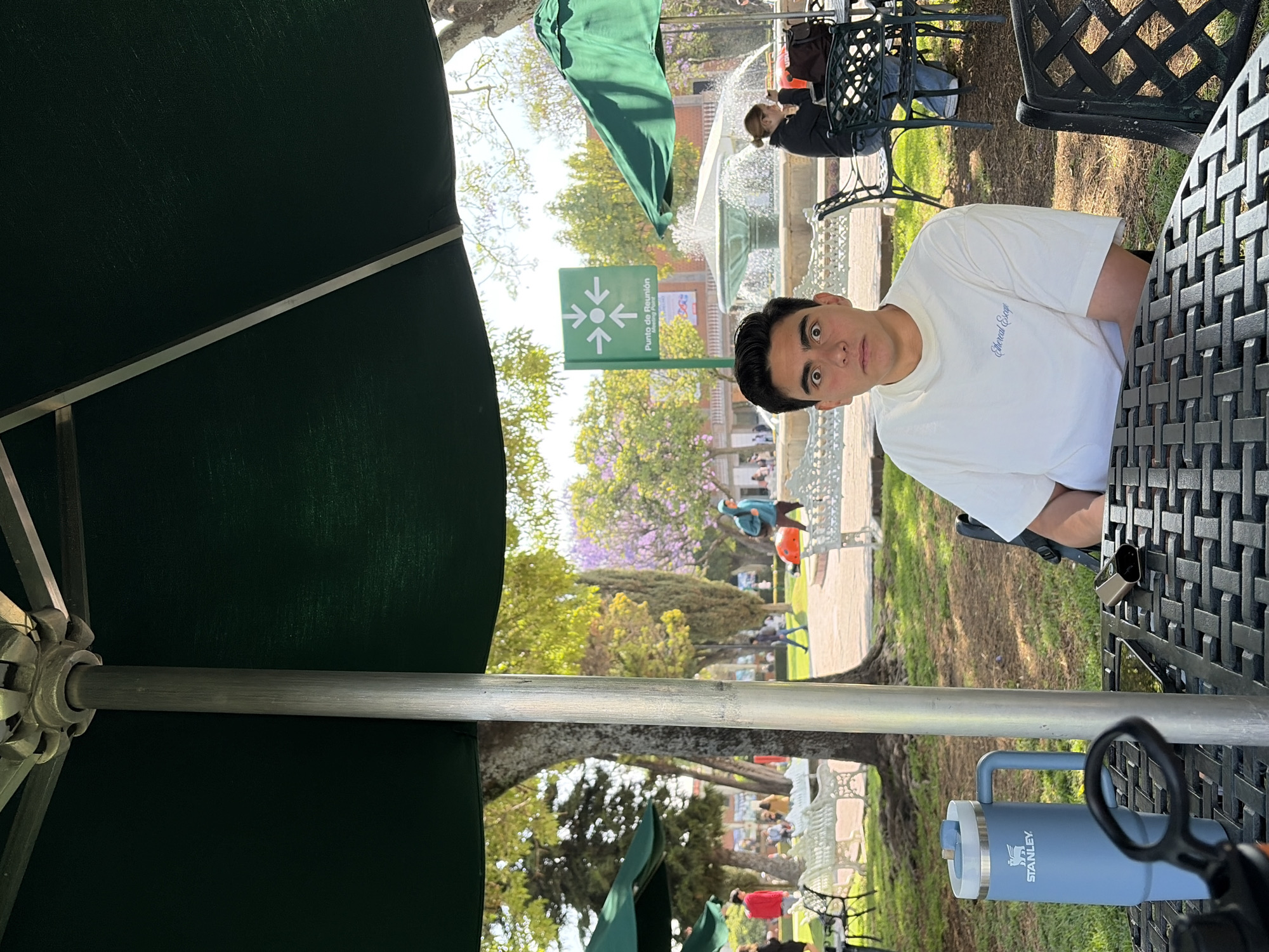 The image shows a young man seated at a black wrought-iron table under a green umbrella. He is wearing a white t-shirt. On the table is a gray Stanley brand water bottle, a phone, and a small black object. In the background, there is a water fountain, a green sign that reads "Punto de Reunión" with arrows pointing inward, and trees with purple flowers. People are walking in the background. The location appears to be in the city of San Andrés Cholula, Mexico.