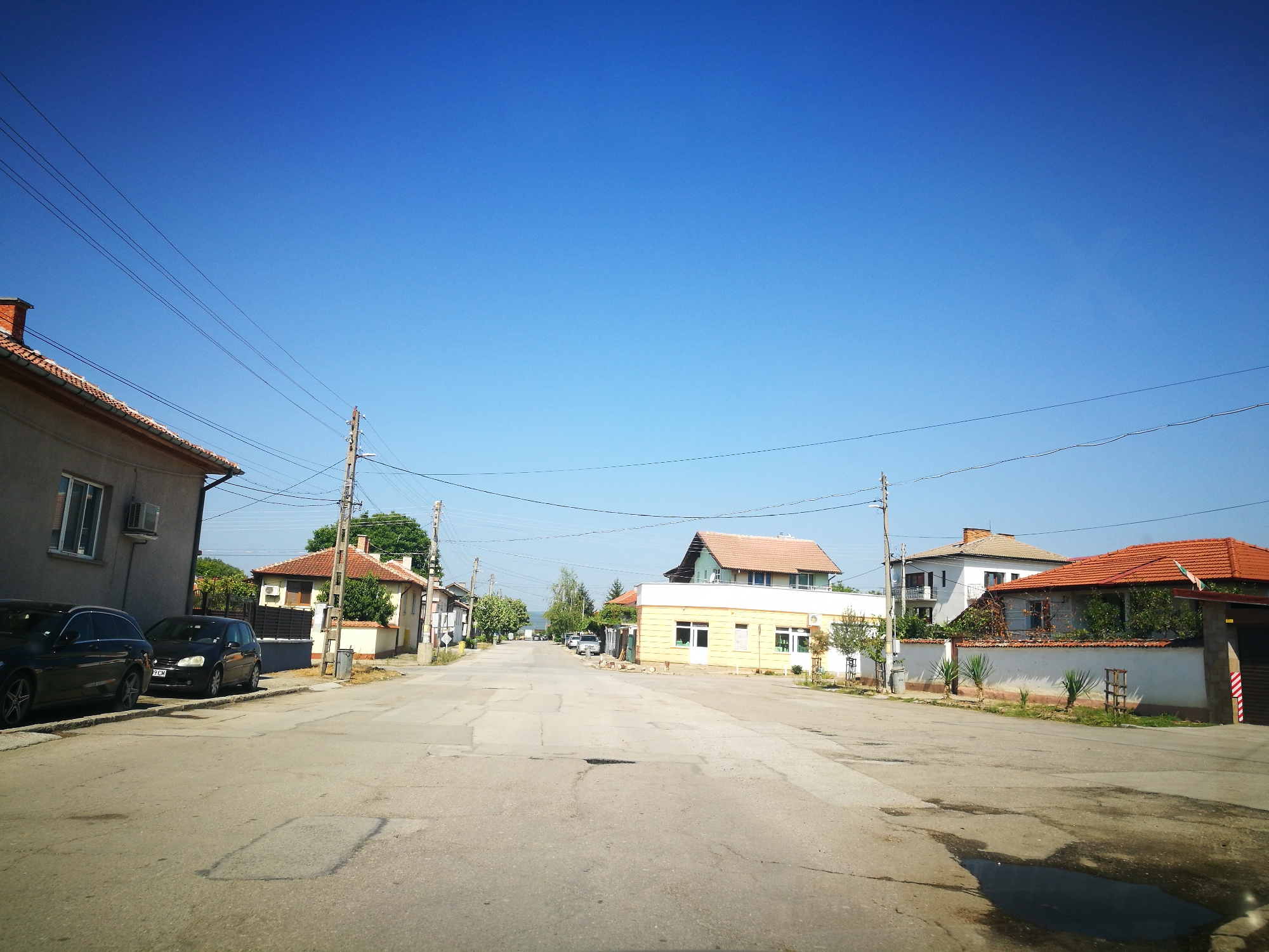 This is a daytime, eye-level, outdoor, full shot image of a street scene in Ihtiman, Bulgaria. The sky is a clear, bright blue with no clouds. A paved road, showing signs of wear and tear with some dark patches and cracks, runs through the center of the frame, extending into the distance. Power lines crisscross the sky above the street, supported by wooden utility poles.
On the left side of the street, a two-story building with a light-colored facade and a dark-tiled roof is visible. An air conditioning unit is mounted on the exterior wall. Parked parallel to the curb are two dark-colored cars. The sidewalk area in front of this building is lined with a low stone wall.
Further down the street on the left, several other houses with tiled roofs are visible, nestled among trees.
On the right side of the street, a yellow, single-story building with large windows and white trim appears to be a commercial establishment. It has a white door and planters with greenery. Adjacent to this building, a white wall topped with an orange-tiled roof forms the boundary of a property. Yucca plants are planted in front of this wall. A dark, garage-like structure with a red and white striped pole is partially visible to the far right. Several more houses with red-tiled roofs and white walls are scattered along the right side of the street, disappearing into the distance.
The overall impression is a quiet, residential street in a Bulgarian town on a sunny day.