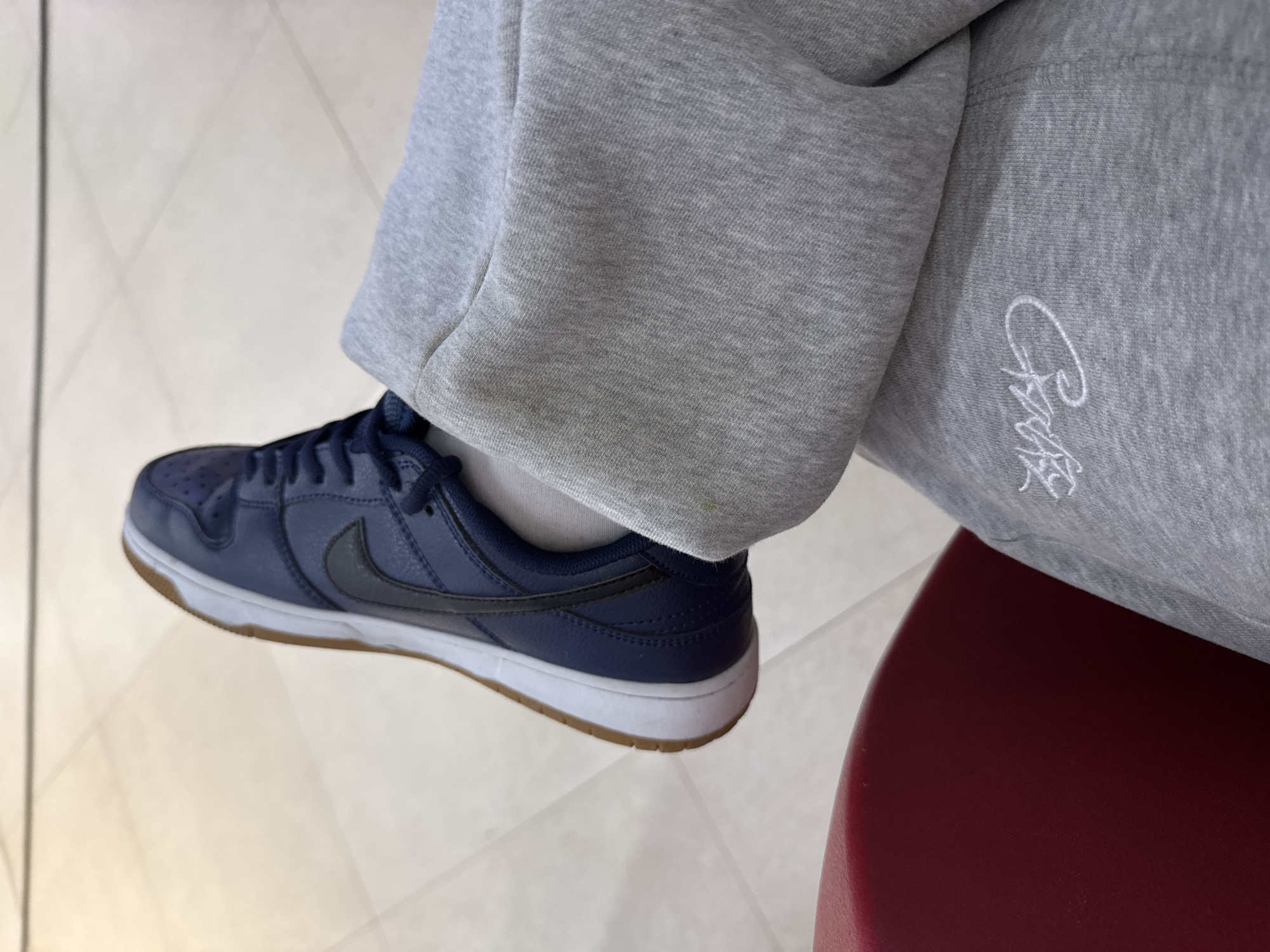 A close-up shot shows a person wearing a navy blue Nike Dunk Low sneaker with a white midsole and brown outsole. The person is also wearing gray sweatpants with a white embroidered design on the right leg. The footwear and leg are positioned on a light-colored tiled floor. The edge of a burgundy-colored chair is visible on the right side of the frame. The image does not provide enough context to determine the specific location in Yerevan, Armenia.