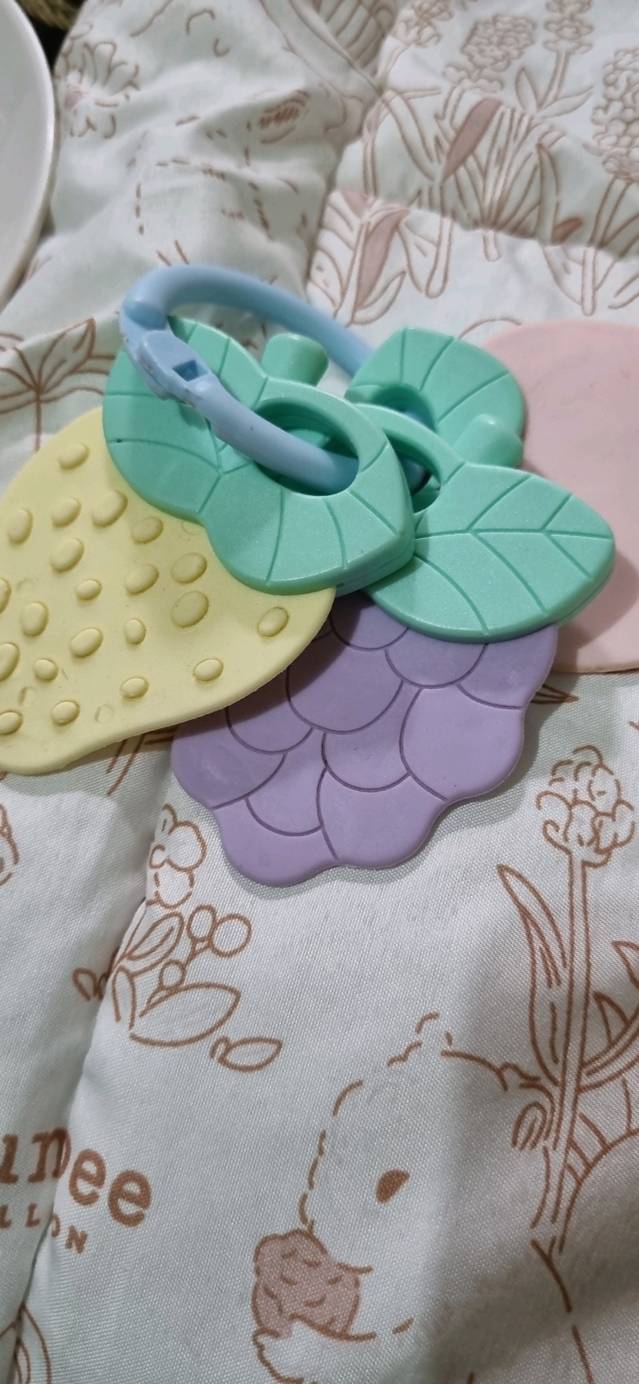 A close-up, top-down shot shows a set of baby teethers resting on a light-colored, patterned fabric. The teethers are made of silicone and have a blue ring connecting several leaf-shaped pieces in pastel green, yellow, and lavender. The yellow teether has raised circular bumps, and the lavender teether has a scale-like pattern. The fabric beneath has a subtle floral or botanical print in a light brown hue, and part of a white bowl is visible on the left edge. There are no people or actions depicted, and the scene is static. The image is well-lit, and the focus is sharp on the teethers. The context of Denpasar, Indonesia is not visually determinable from the provided image.